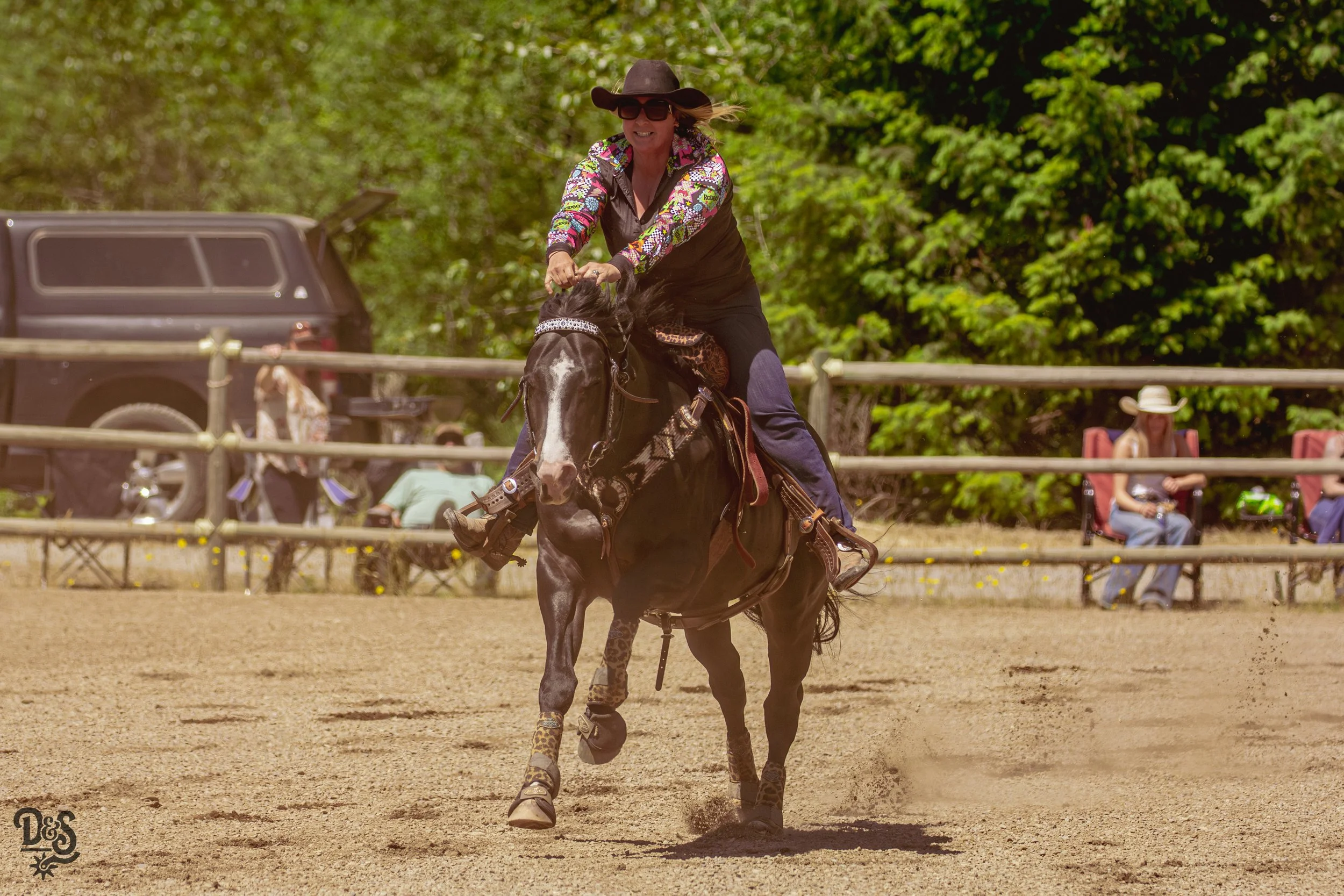 A woman in a cowboy hat, sunglasses, and a colorful long-sleeve shirt rides a horse in an outdoor arena with green trees and spectators in chairs in the background.