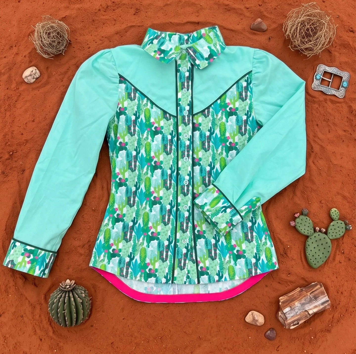 A turquoise long-sleeve button-up shirt with cactus patterns, laid flat on reddish soil, surrounded by small desert-themed objects including cacti, rocks, a pinecone, and a rhinestone belt buckle.