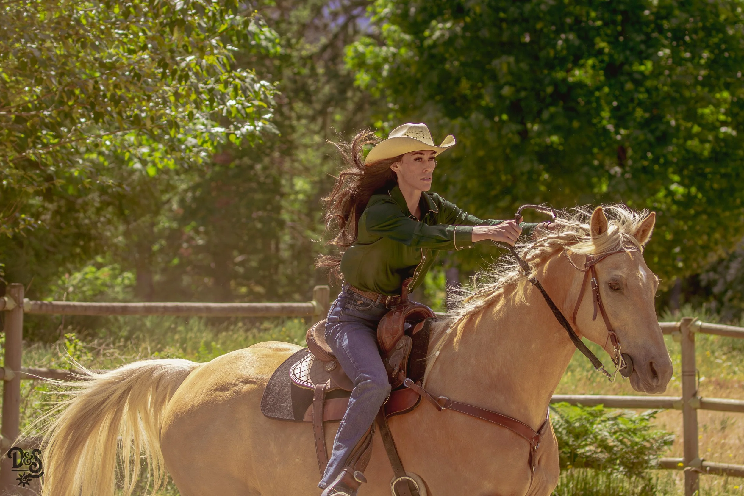 A woman wearing a cowboy hat and green shirt riding a light-colored horse through a wooded area.