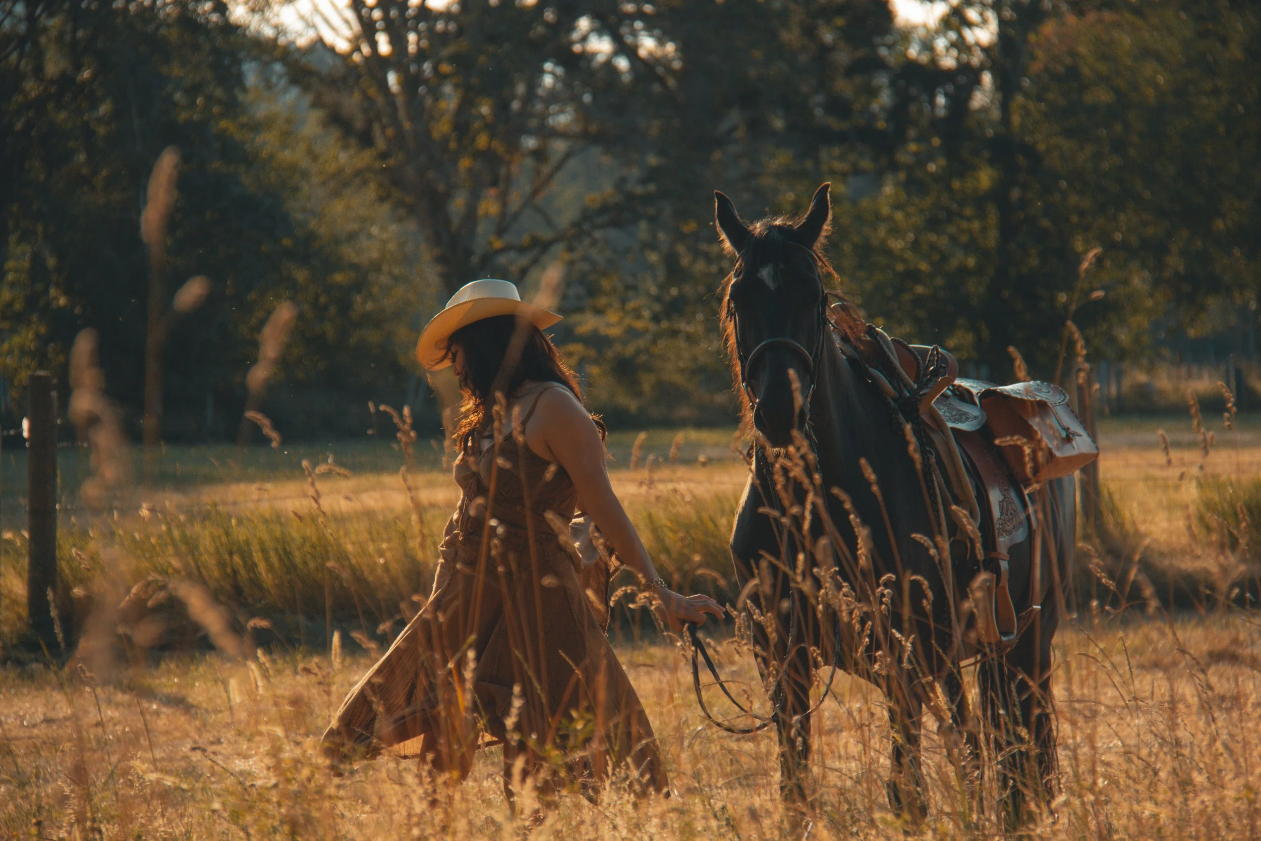 A person wearing a hat and long dress walking through tall grass with a horse leading the way in a forested area during sunset.