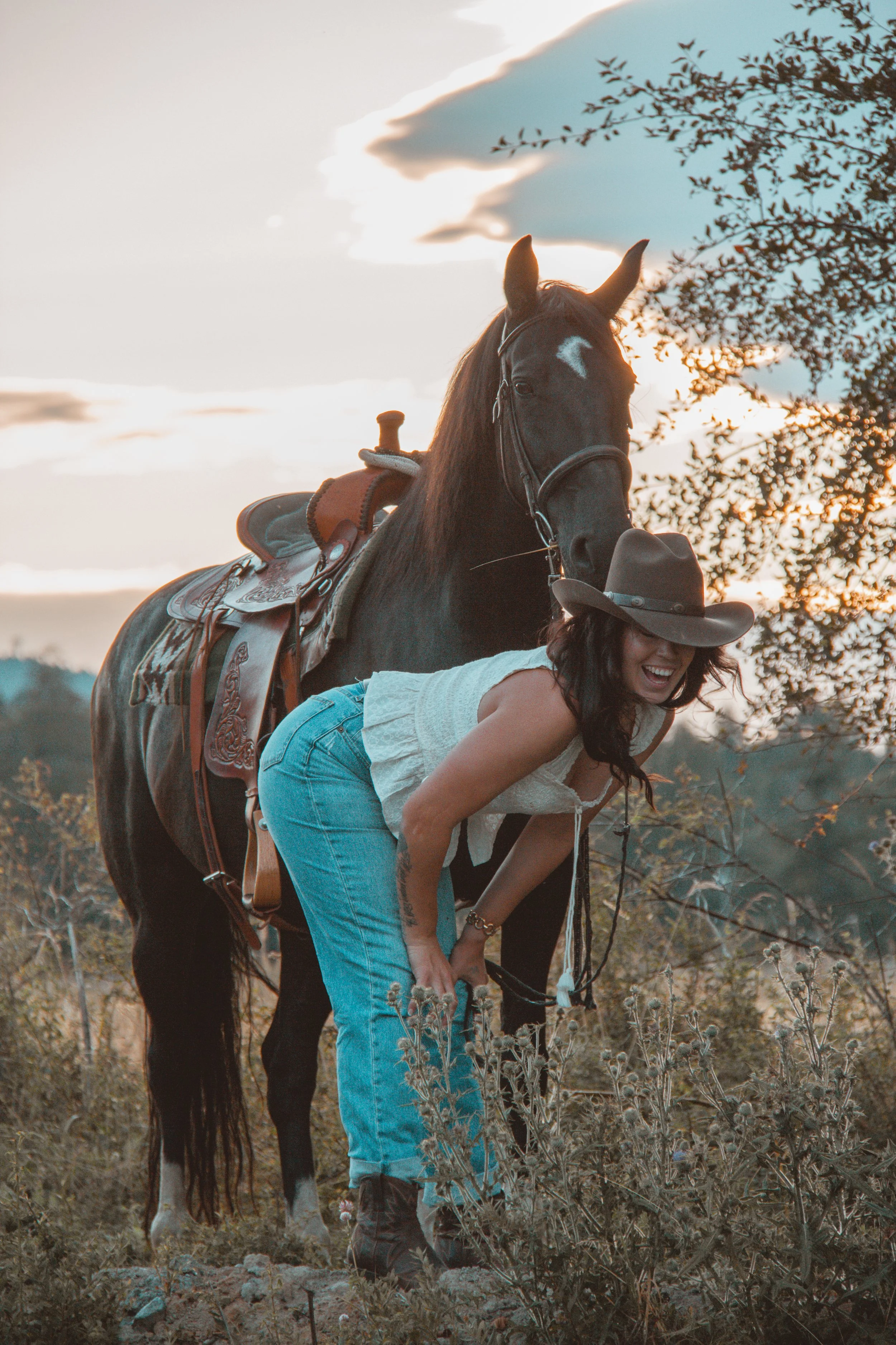 A woman in a white sleeveless top, jeans, and a cowboy hat is bending over, riding a black horse with a decorative saddle, in an outdoor natural setting during sunset. The horse's head is raised and facing forward, while the woman is smiling and look