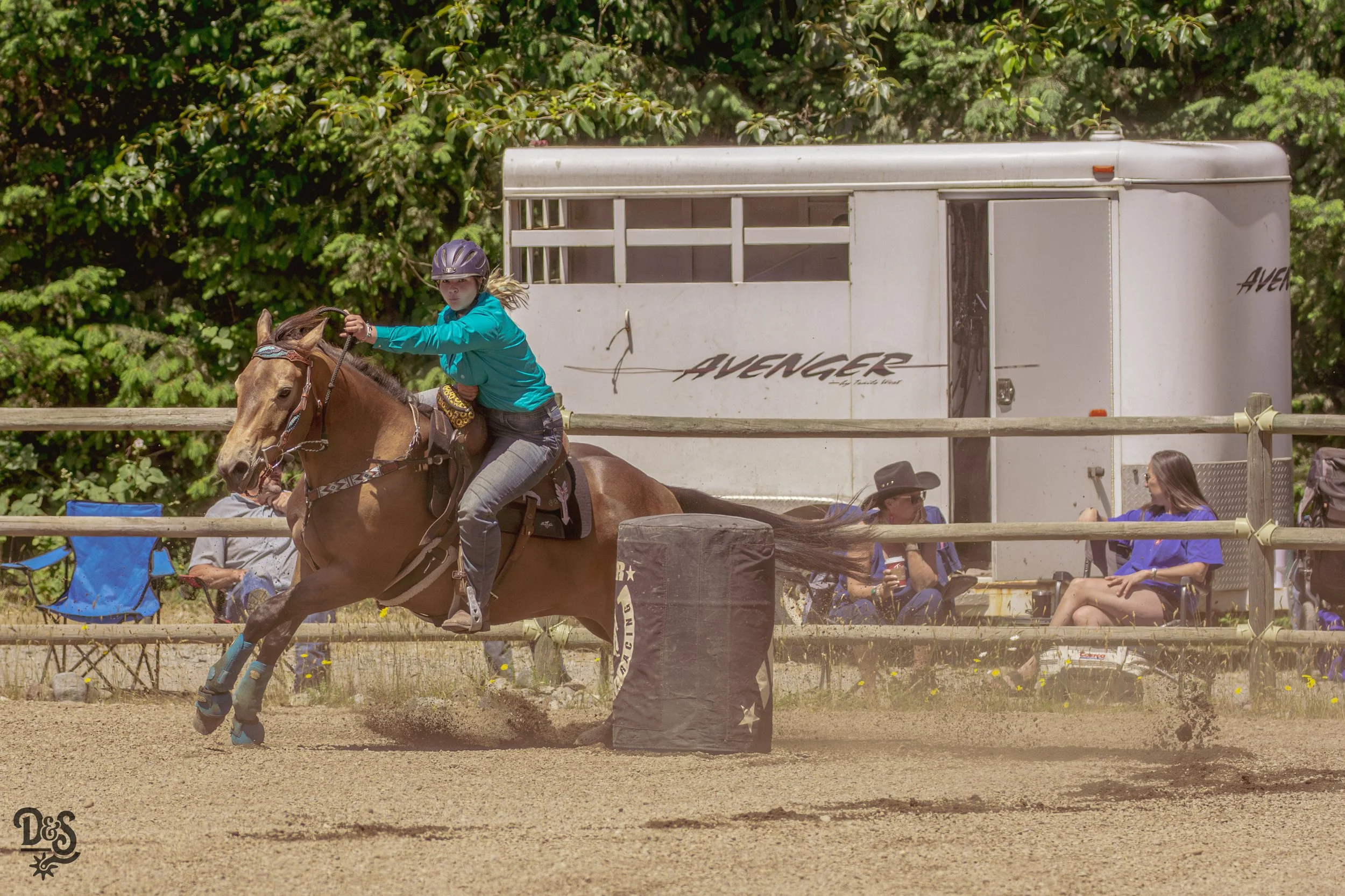 A young girl wearing a helmet rides a horse in a fenced outdoor arena with a trailer and spectators in the background.