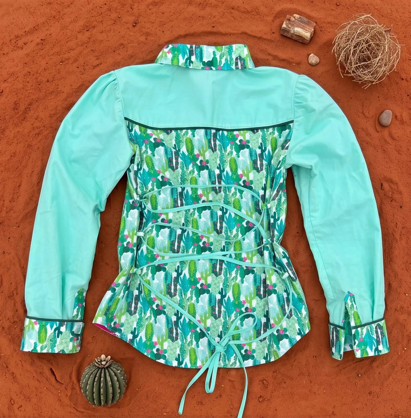 A shirt with a cactus and desert pattern lies on red desert sand surrounded by small rocks and a round cactus plant.