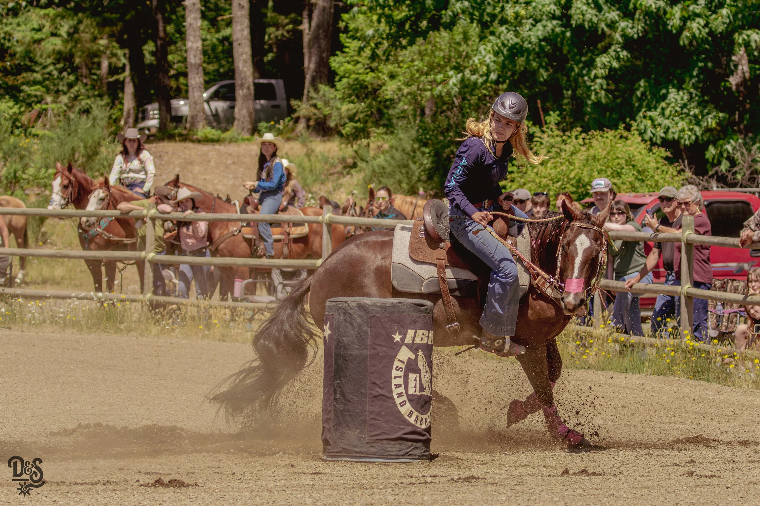 A young woman wearing a helmet riding a horse in an outdoor arena, with spectators and trees in the background.