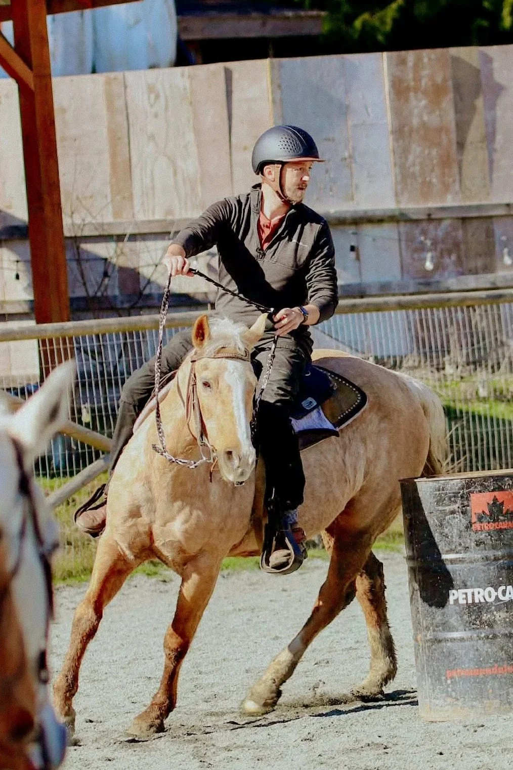 Man wearing a helmet riding a light-colored horse in an outdoor riding arena, with a barrel and fencing visible in the background.