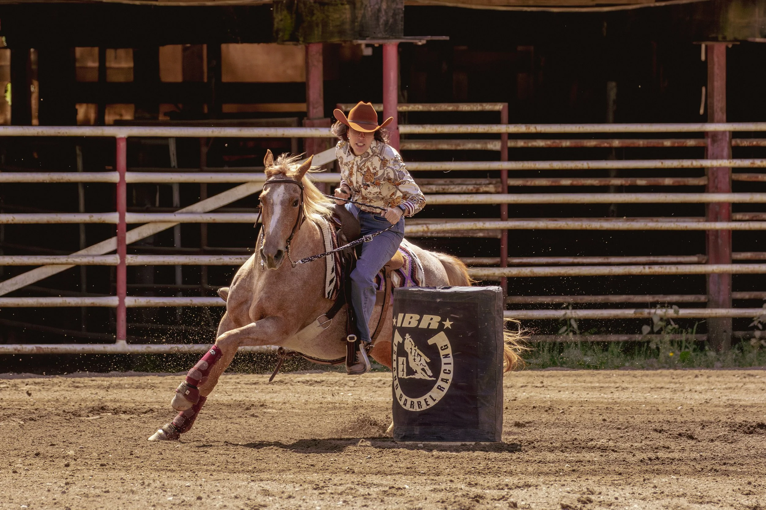 Rodeo rider on a horse with leg wraps, wearing a cowboy hat, jeans, and a patterned shirt, approaching a barrel marked 'RACING' on an outdoor dirt arena.