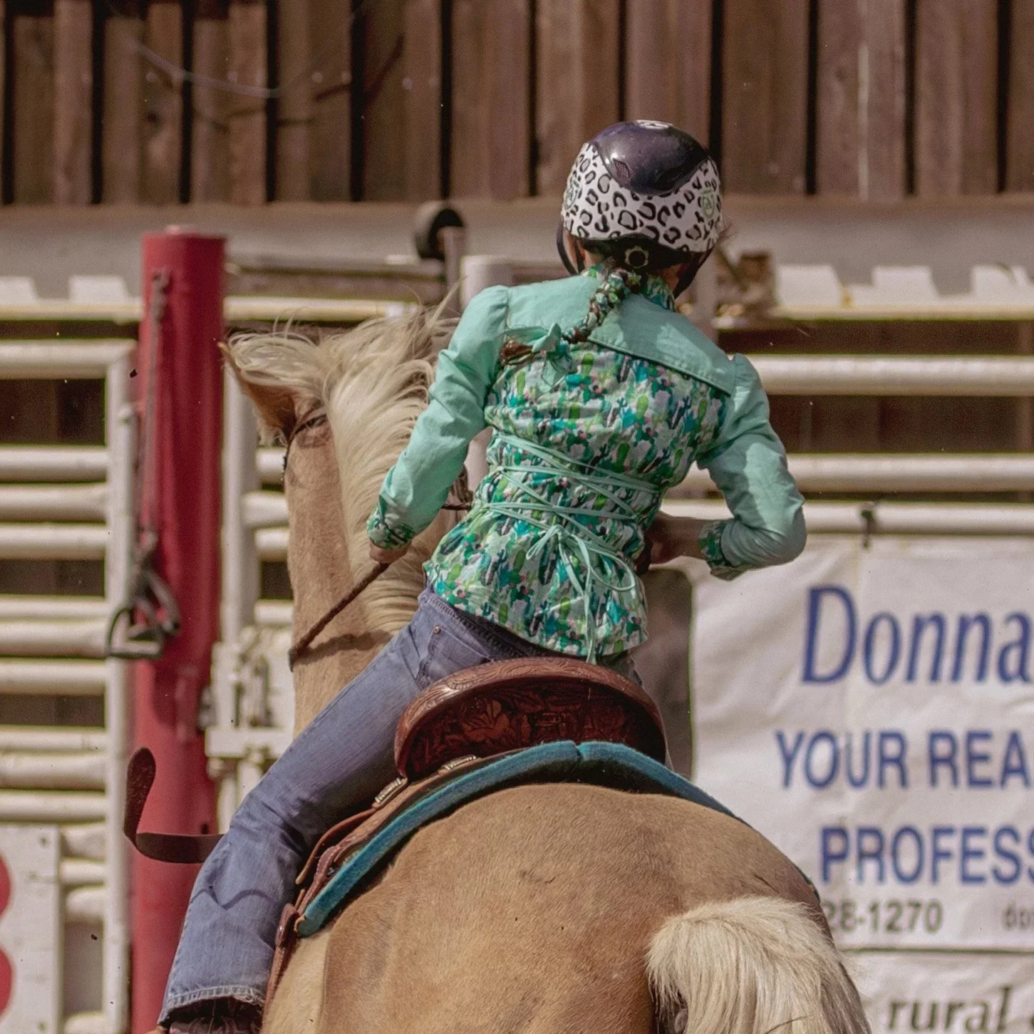 A person wearing a helmet and a green patterned jacket riding a horse in an indoor arena.