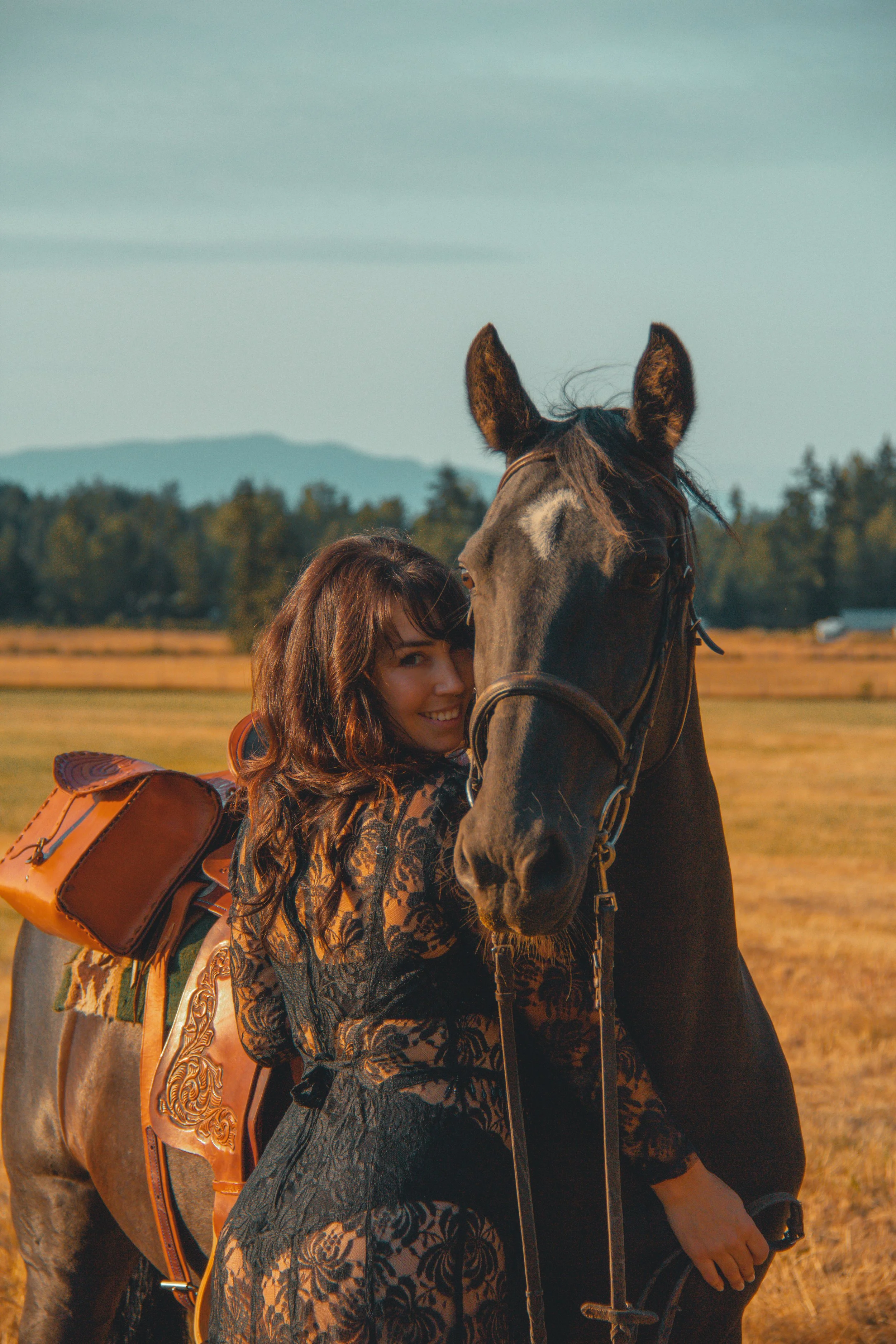A woman with long, wavy brown hair and a patterned black dress is standing next to a large, dark horse with a white star on its forehead. The woman is smiling and leaning her head against the horse. The woman has a large, decorative leather saddle ba