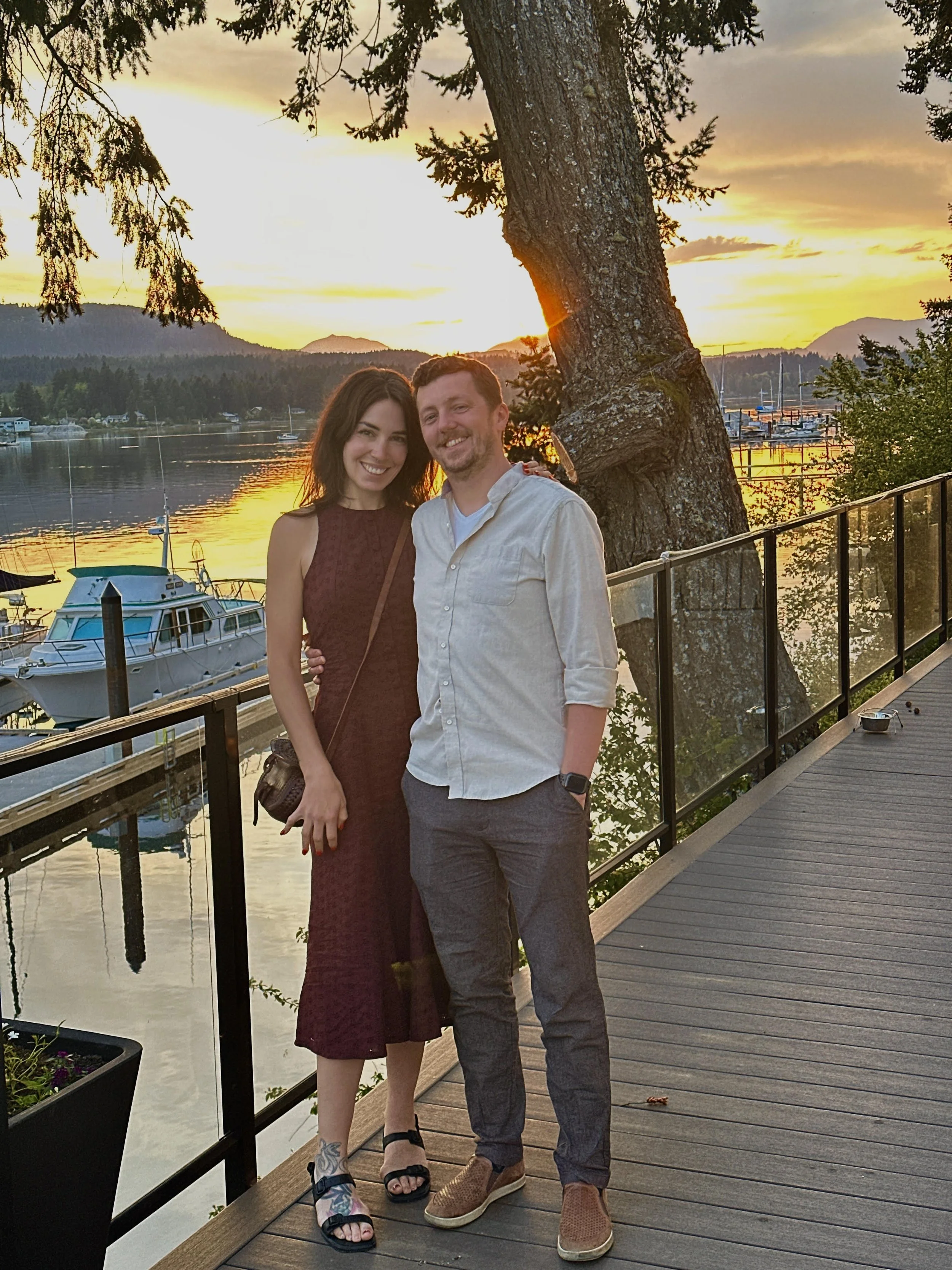 A smiling couple standing on a wooden dock near a marina during sunset, with boats docked in the water and trees in the background.