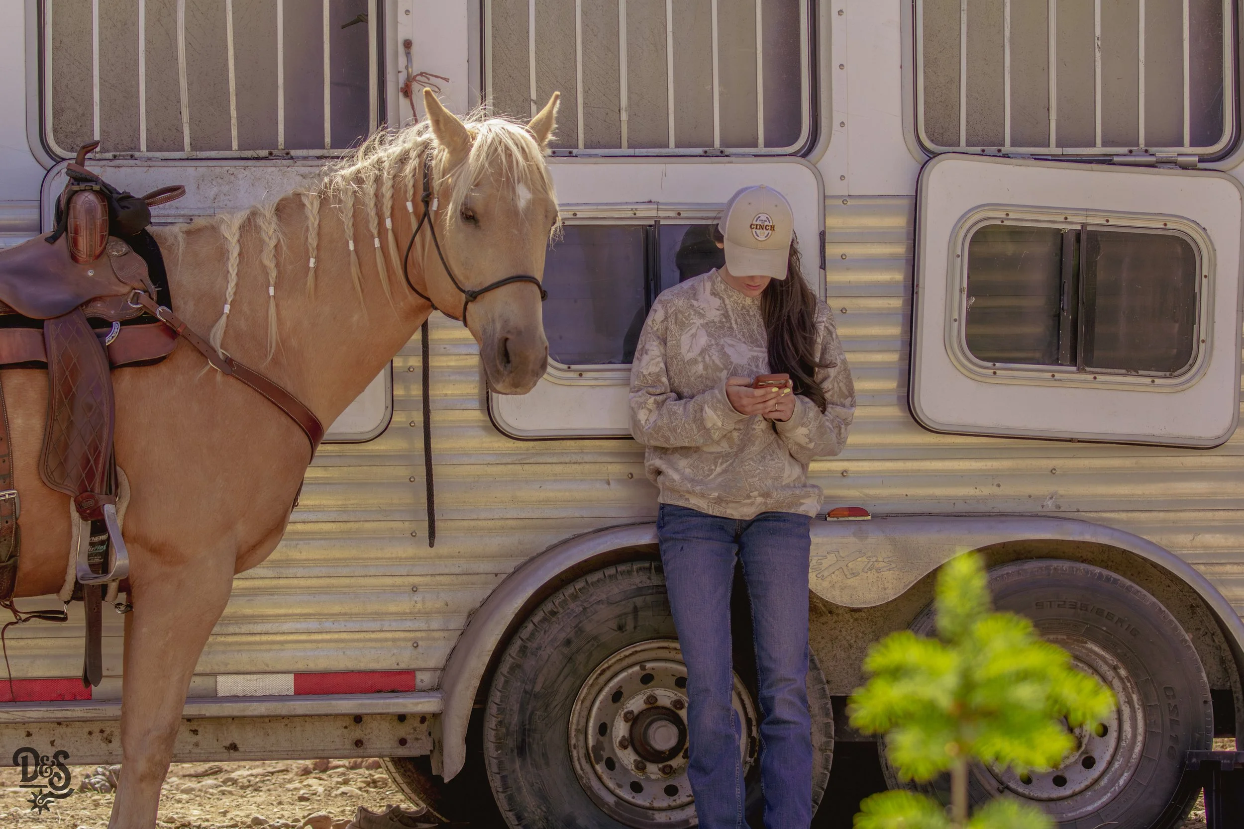 A woman in a baseball cap using her phone leans against a horse trailer with a saddled horse beside her.