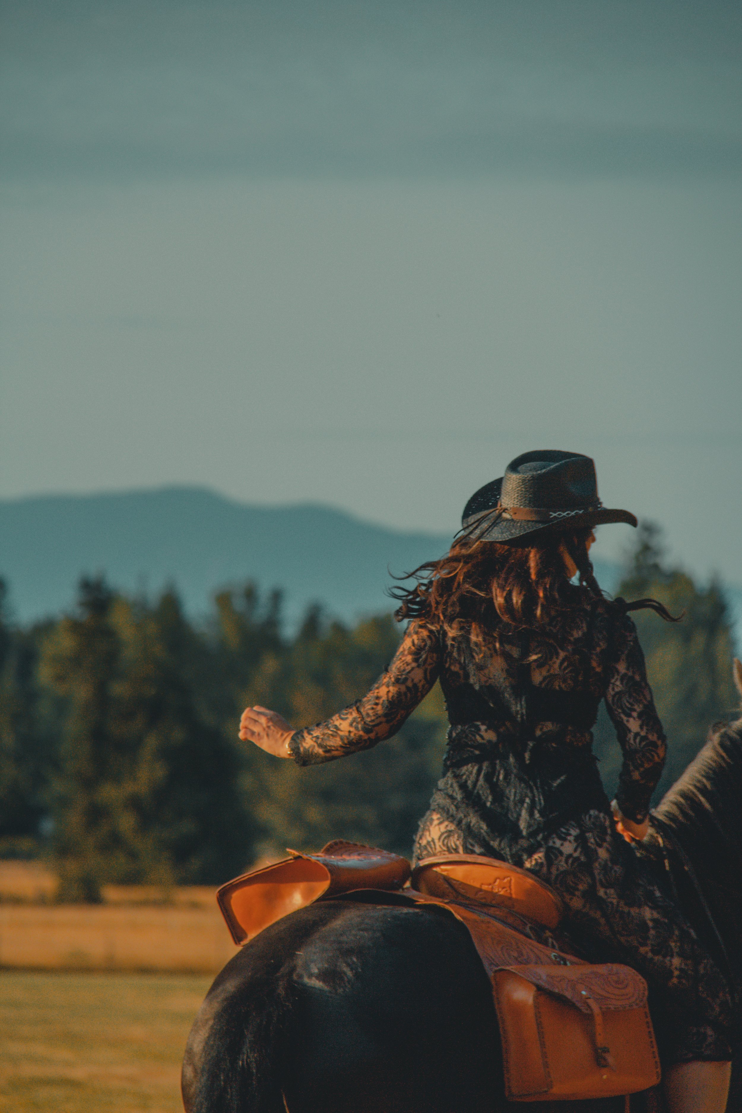 A woman with long curly hair wearing a cowboy hat and a black lace top riding a horse in a rural landscape during sunset.