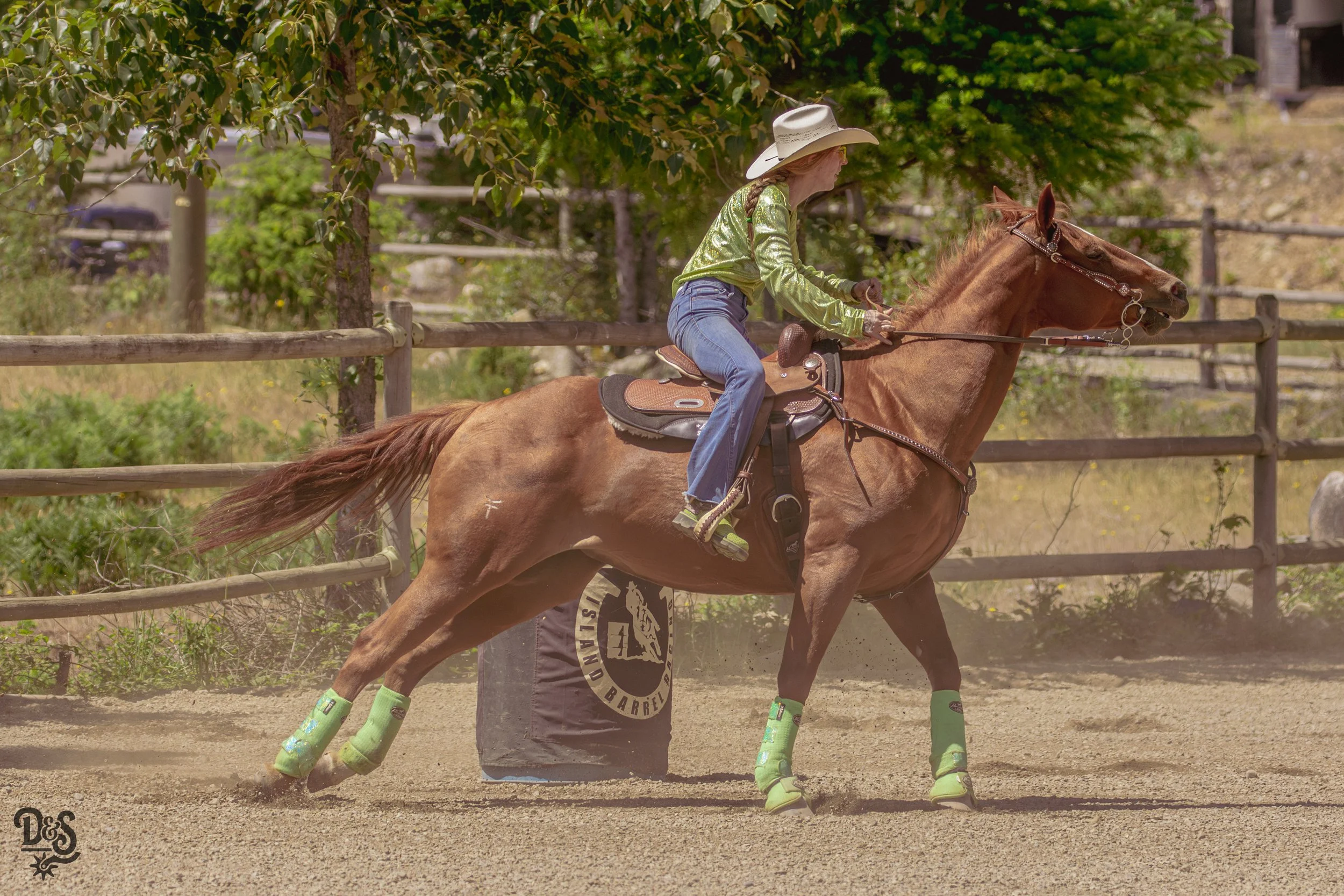 A woman riding a chestnut horse on a dirt track, wearing a white cowboy hat, a shiny green jacket, blue jeans, and green leg wraps.