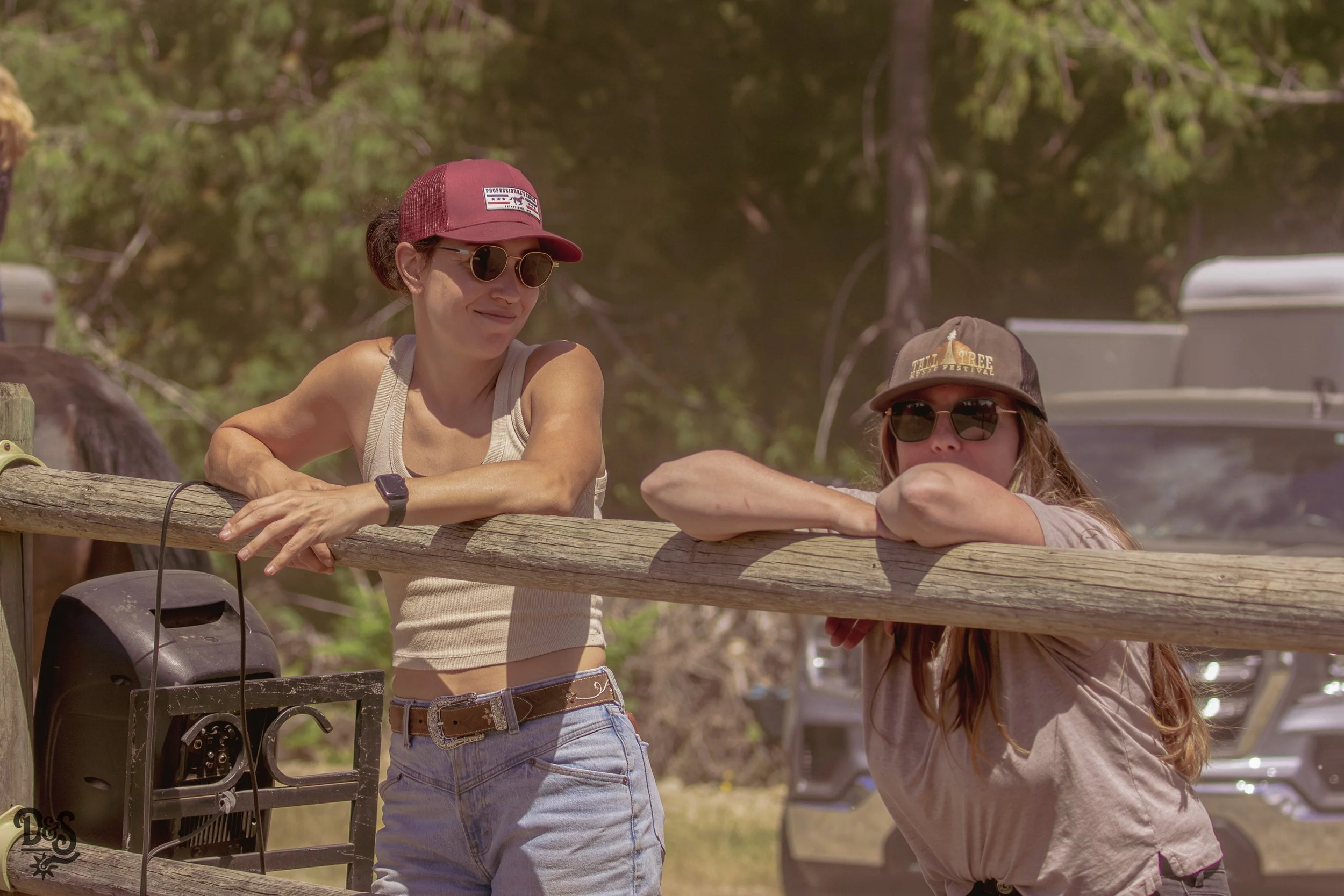 Two women with sunglasses and hats leaning on a wooden railing outdoors, with trees in the background.