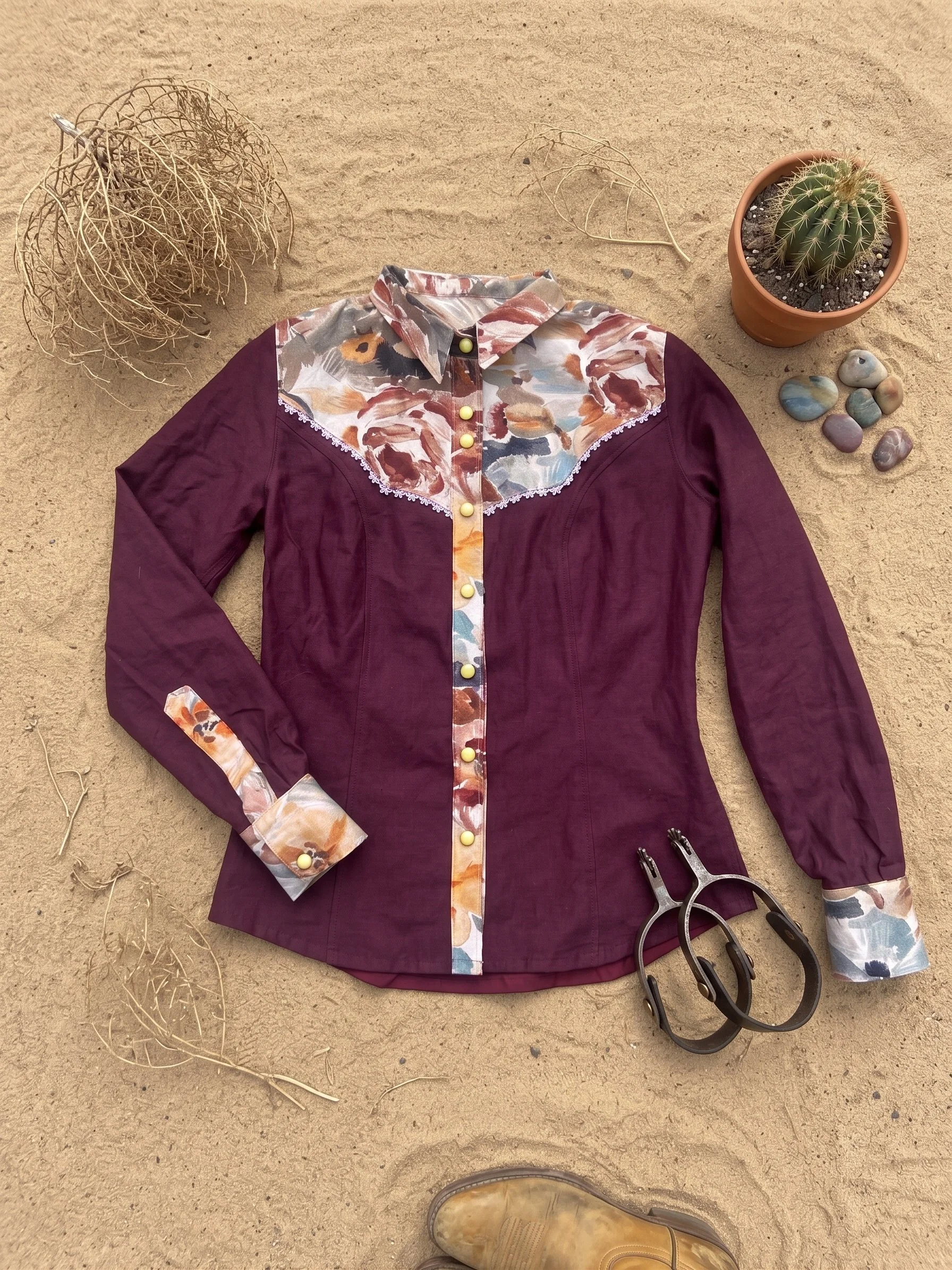 A maroon long-sleeve shirt with floral-patterned collar, cuffs, and front placket, lying on sandy ground with a pair of black-handled pliers, a potted cactus, a dried plant, some small stones, and the toe of a tan boot visible at the bottom of the image.
