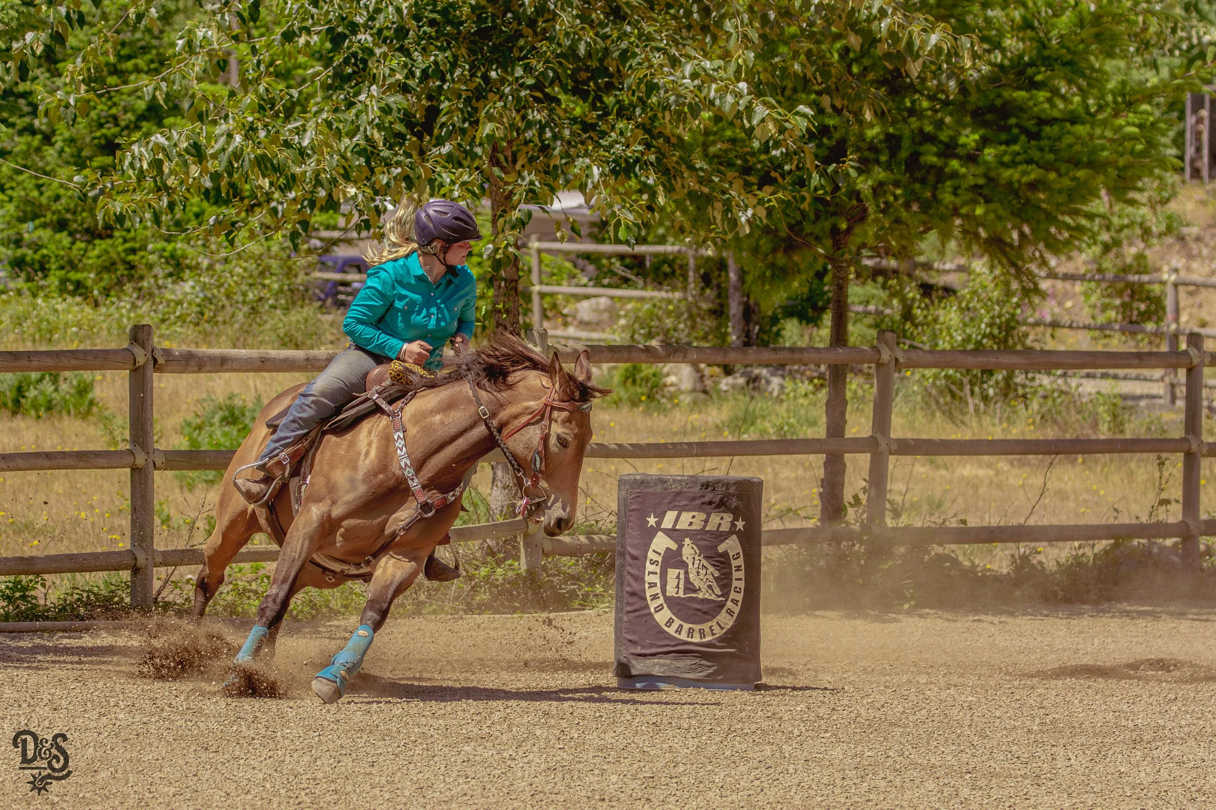 Woman riding a horse during a barrel racing event on a dirt track, wearing a helmet and turquoise shirt, with trees in the background.