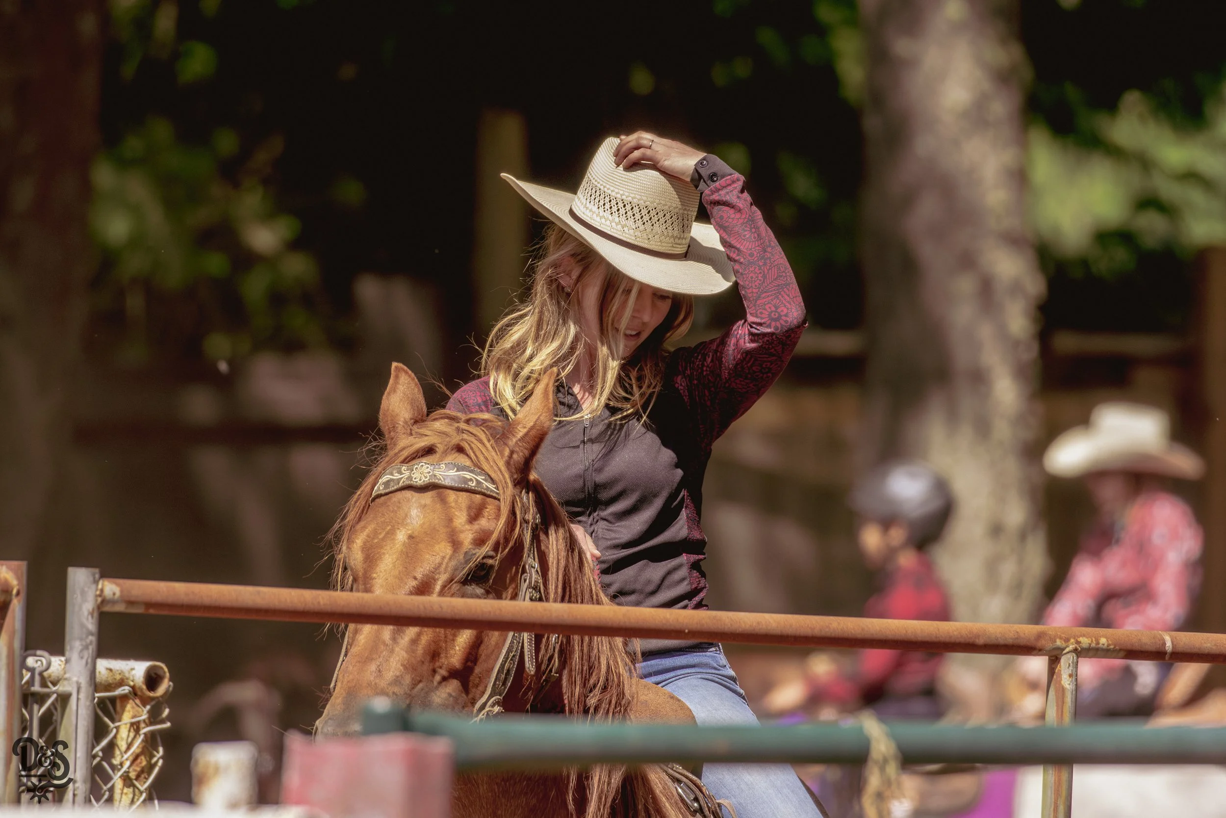 A woman with wavy blonde hair wearing a wide-brimmed straw hat and a dark jacket, sitting on a brown horse, outdoors with trees in the background.