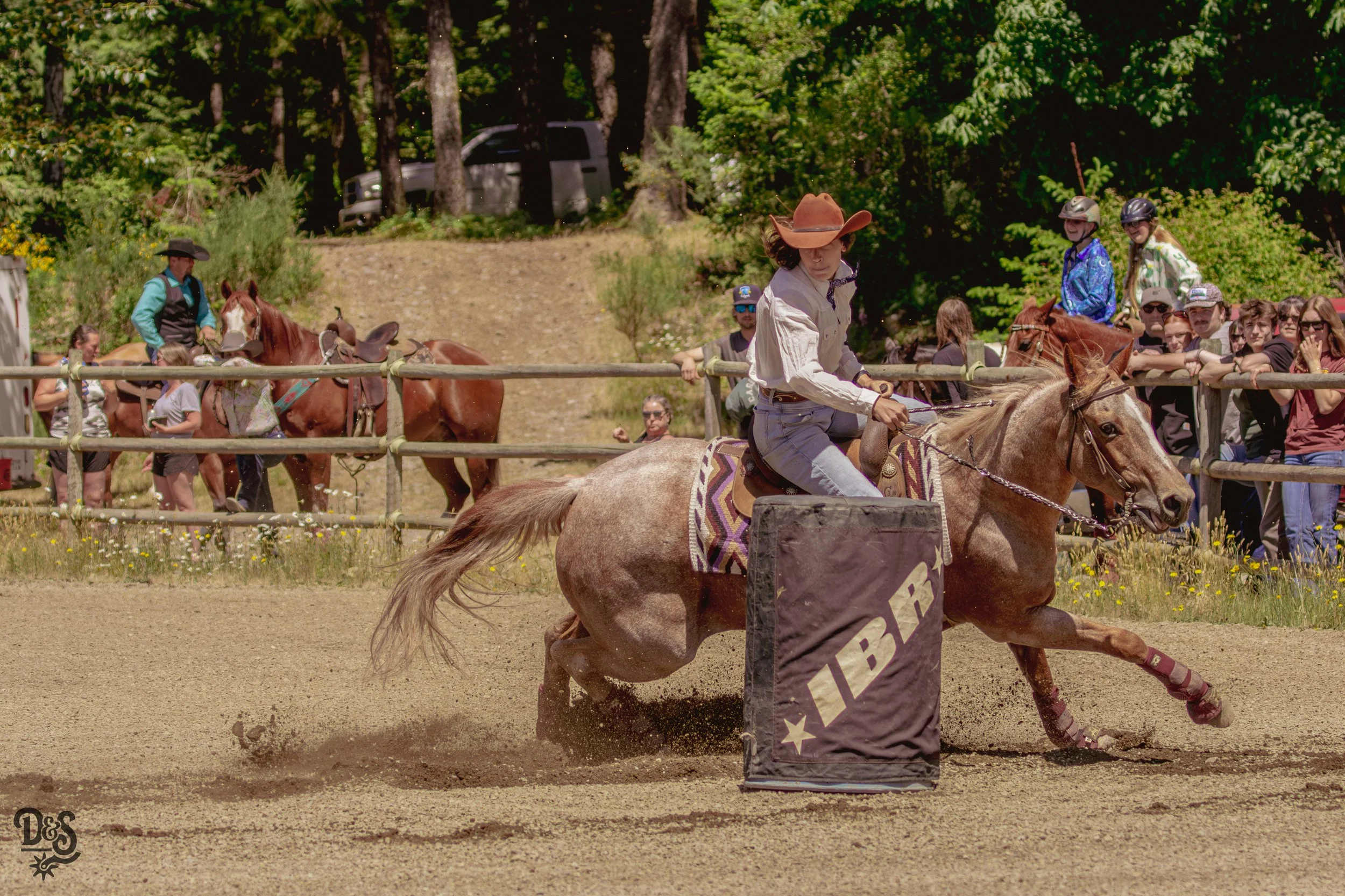 A person riding a horse in a rodeo arena with spectators lined along the fence, trees in the background, and other riders preparing on horses.