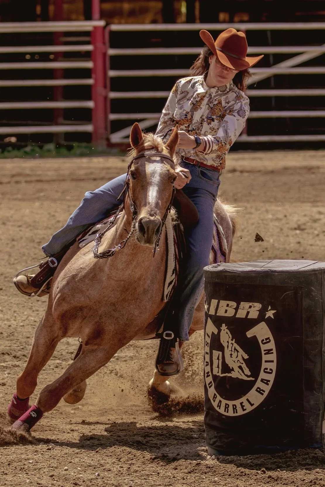 A woman riding a light brown horse during barrel racing. She is wearing a cowboy hat, glasses, a patterned shirt, jeans, and boots. The horse is hitting a barrel marked with the logo 'NBRX Barrel Racing' on a dirt arena with a wooden fence in the background.