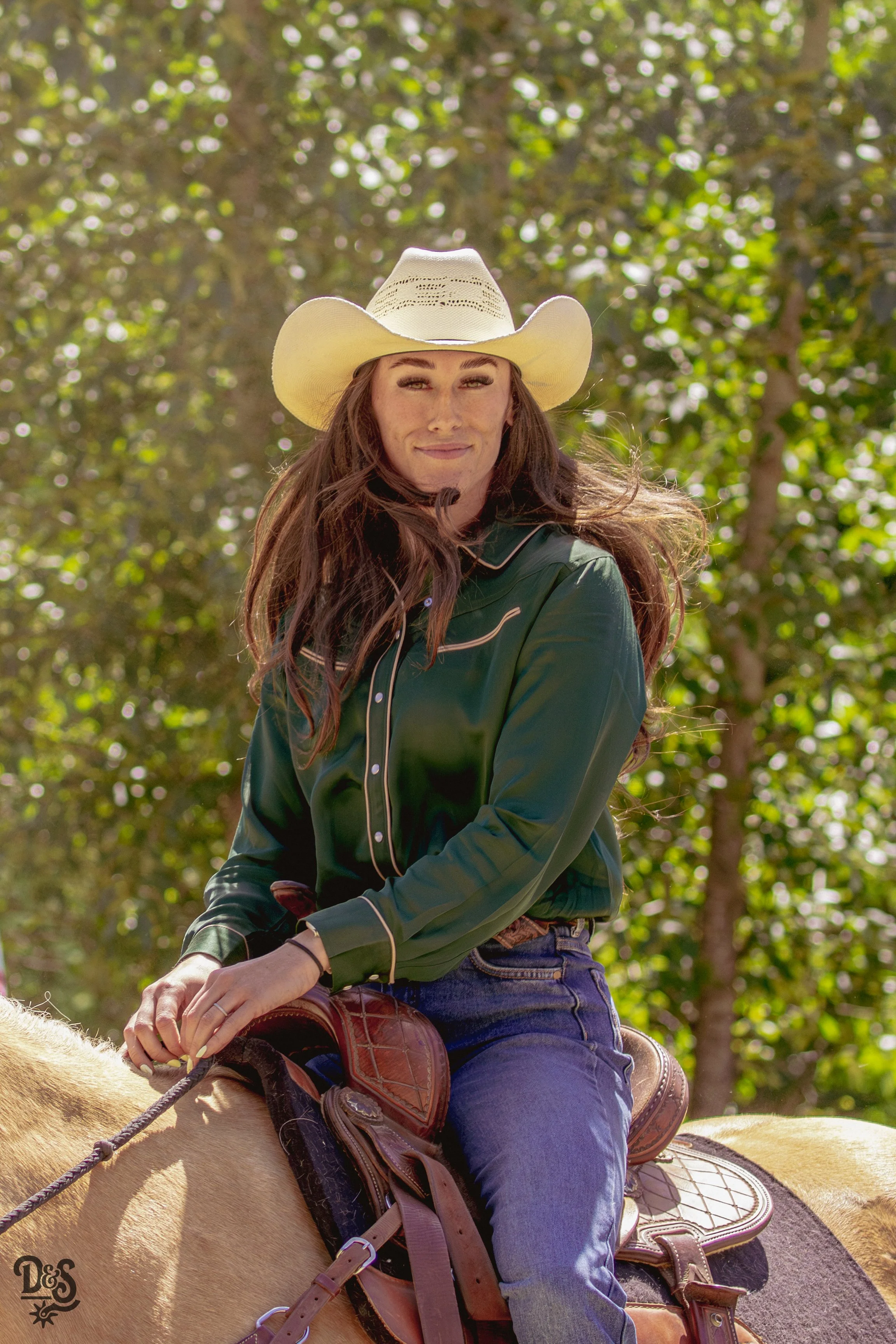 A woman with long brown hair wearing a cowboy hat, green western shirt, jeans, and riding a horse with a saddle, outdoors on a sunny day with green trees in the background.