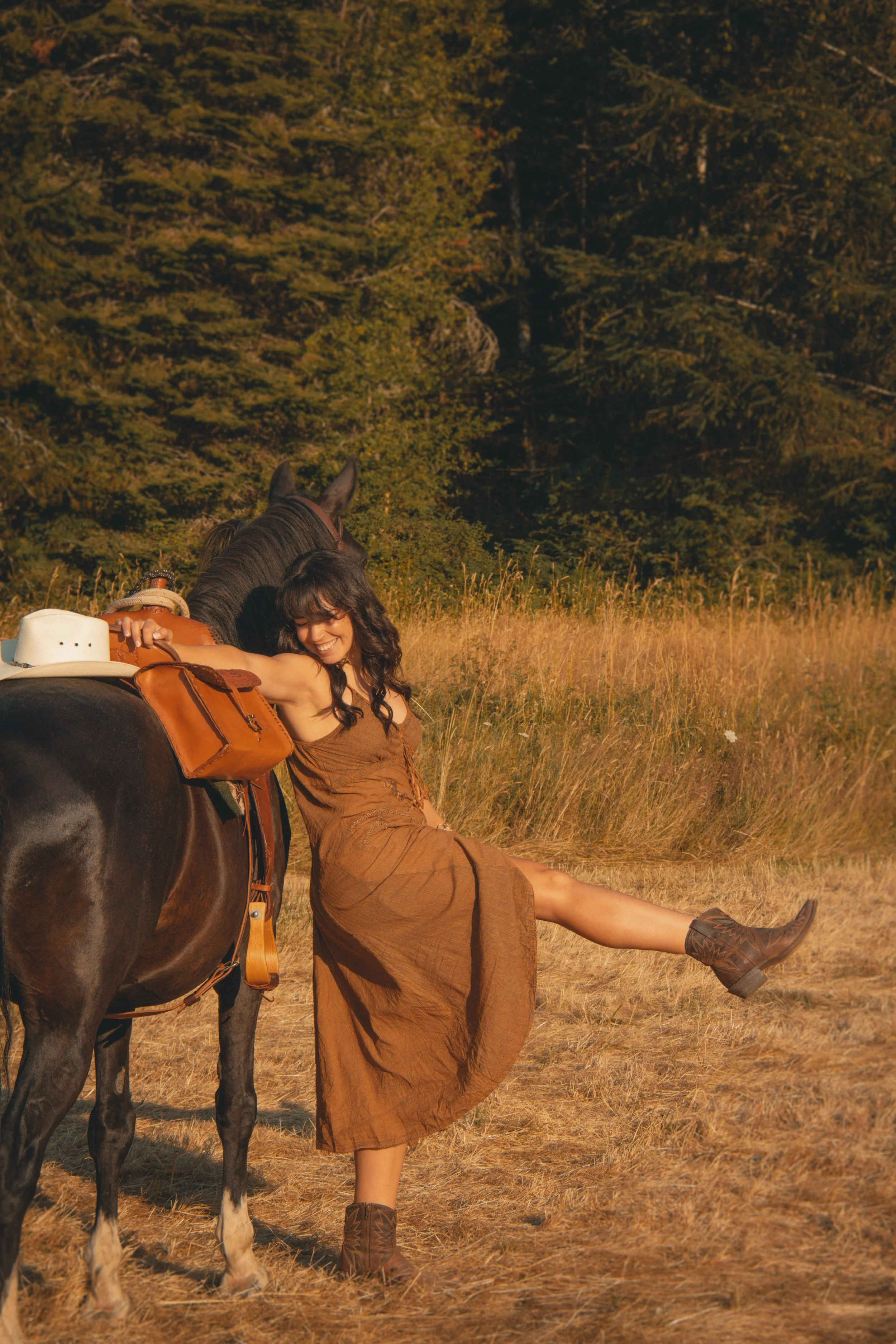 A woman sitting on the ground in a field, leaning on a horse with her eyes closed and a smile, wearing a brown dress and cowboy boots, with a cowboy hat and saddlebag nearby, and a forest in the background during sunset.