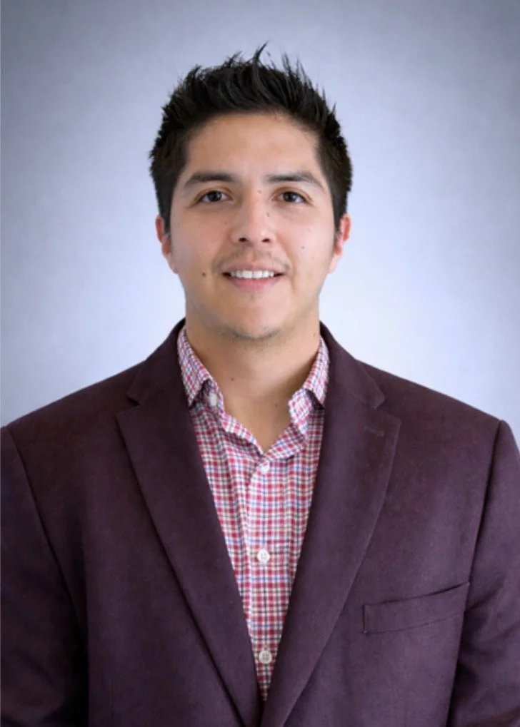 Portrait of a young man with dark hair wearing a burgundy blazer and a red checkered shirt, smiling against a gray background.