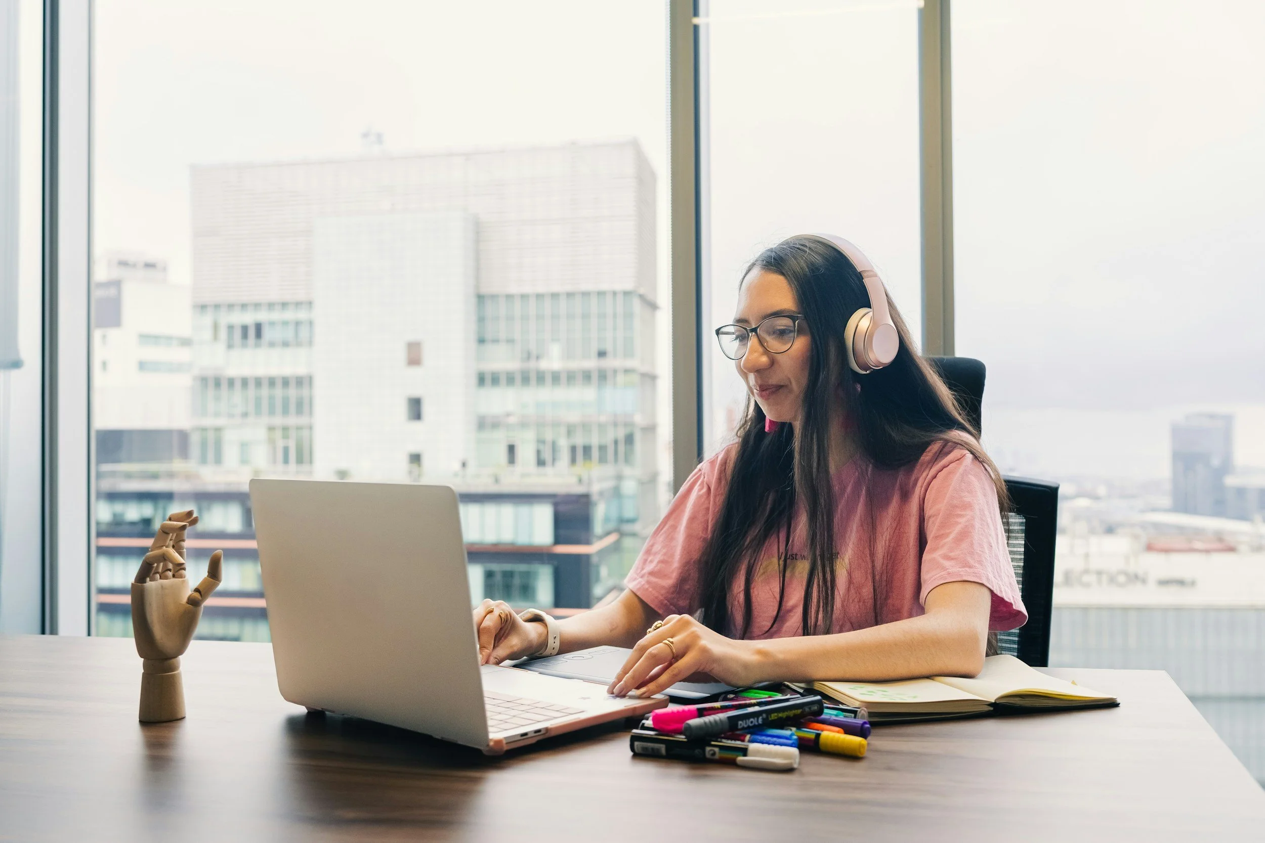 Young woman with long dark hair and glasses working at a desk with a laptop, colorful markers, notebooks, and a wooden mannequin in front of large windows with a cityscape background.