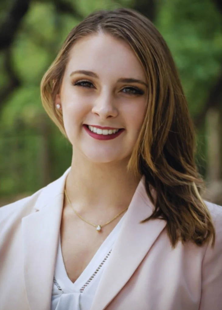 Portrait of a smiling young woman with light brown hair, wearing a light pink blazer and a gold necklace, standing outdoors with greenery in the background.