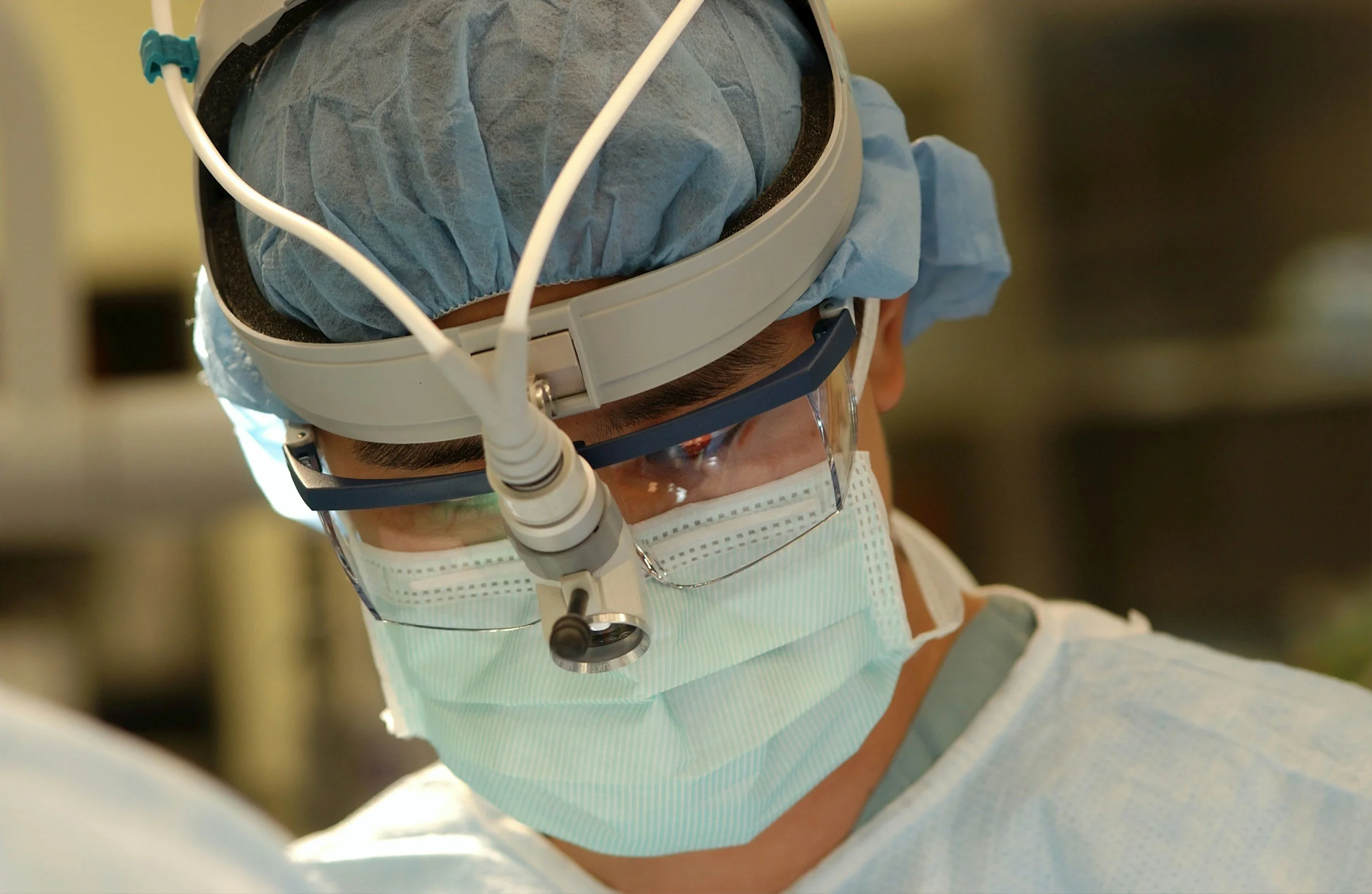 A medical professional wearing a surgical cap, protective glasses, and a face mask, looking downward in a clinical setting.