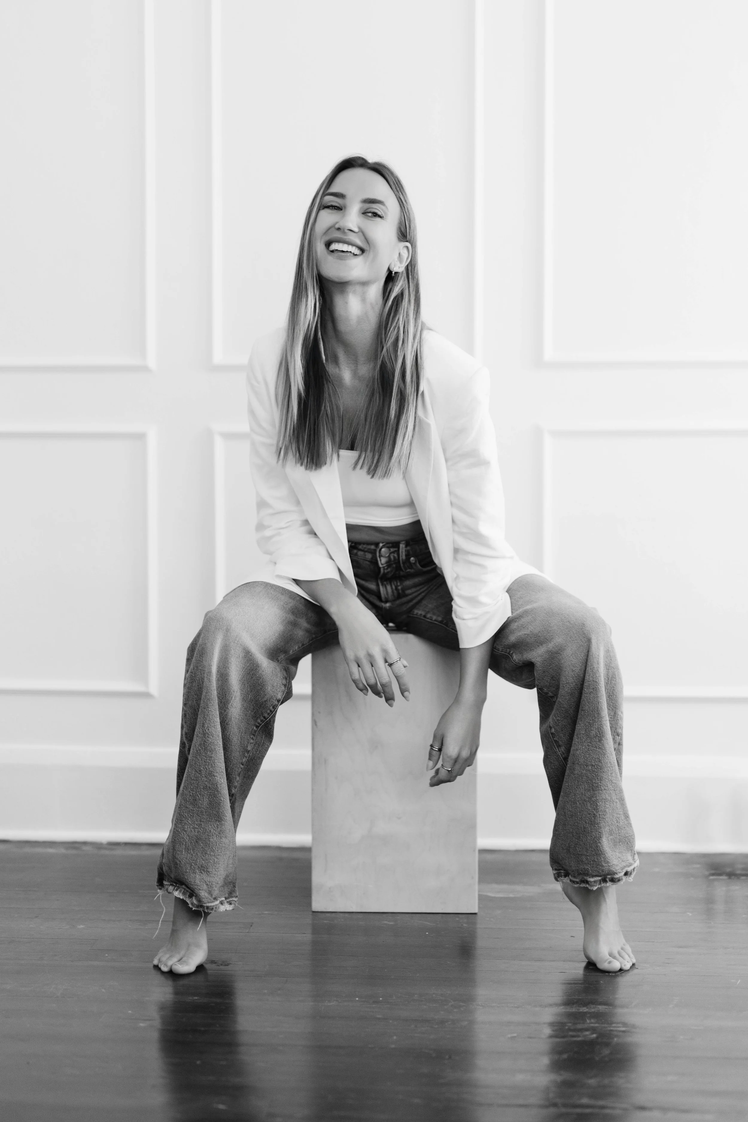 A woman with long hair smiling, sitting on a wooden block with her legs spread apart, feet bare, against a plain white wall with decorative panels, in black and white.
