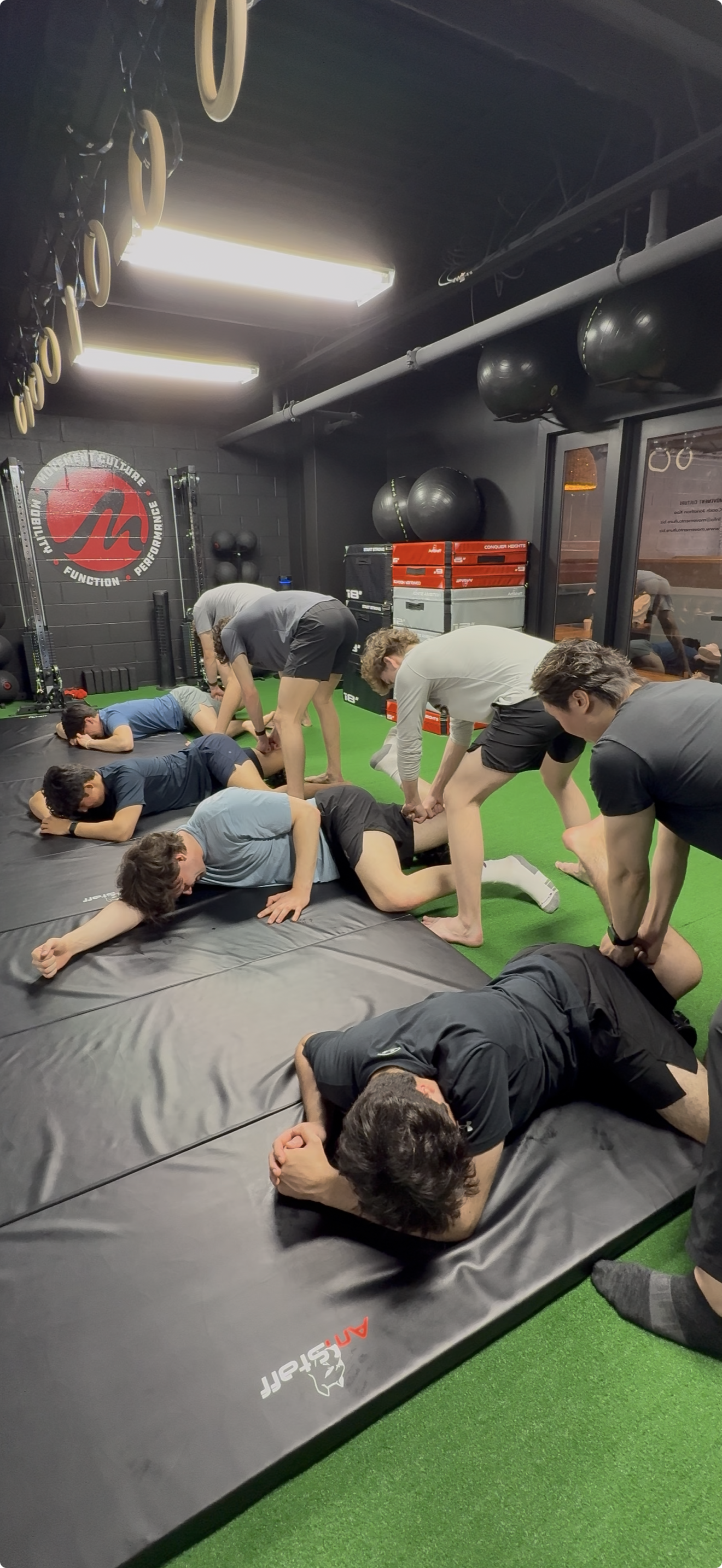 Group of people practicing a wrestling move with some lifting and others on the mat in a gym.