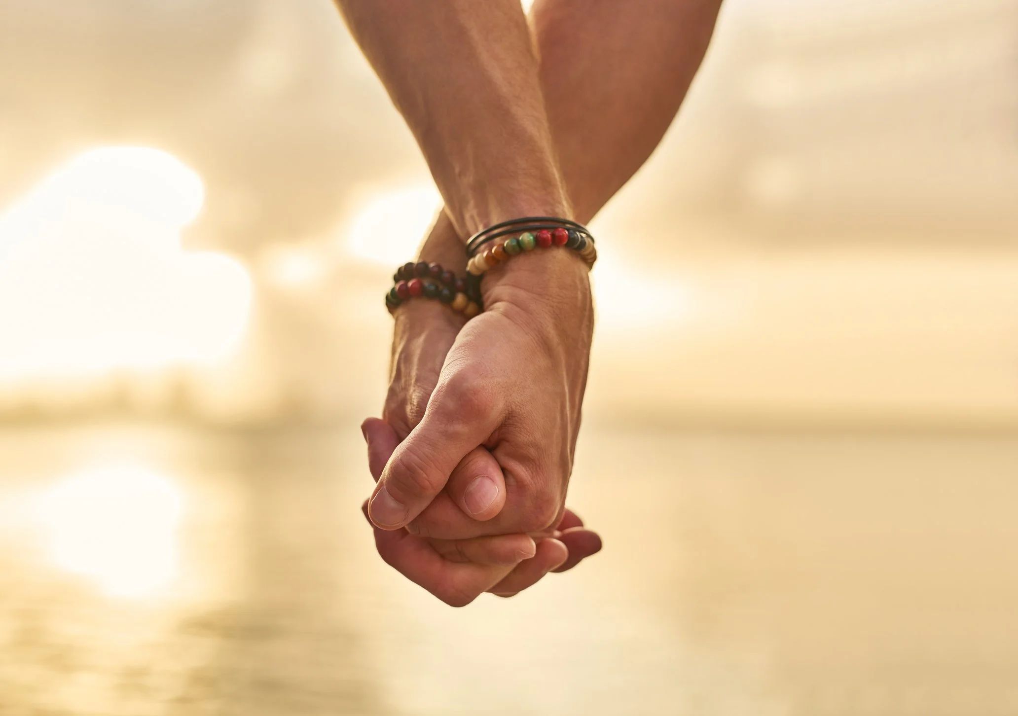 Close-up of two hands clasped together, with several colorful beaded bracelets on one wrist, against a blurred, warm-toned background.