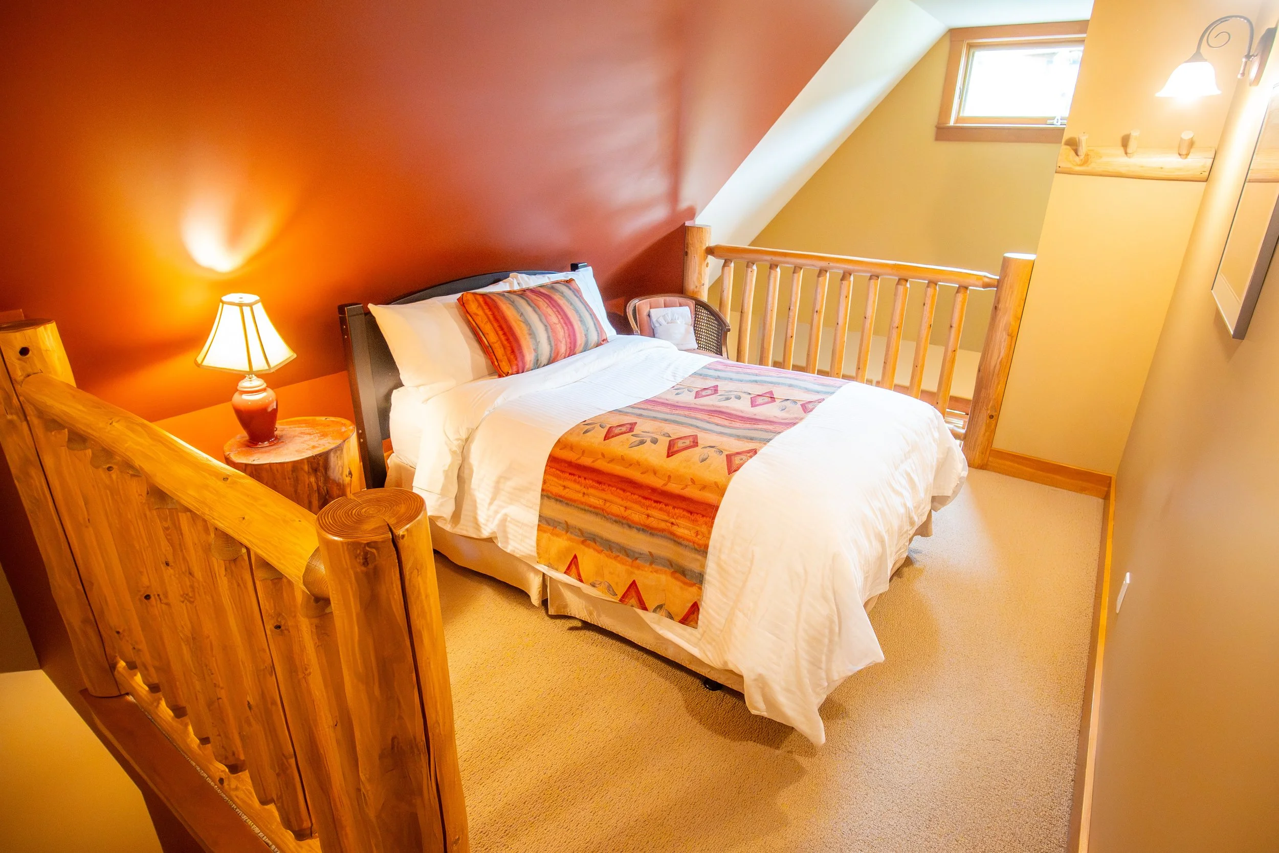 A cozy bedroom with a bed covered in white linens and plaid bedding. There are two folded towels and a towel shaped like a swan on the bed. The room has tan walls, wooden window frames, and blinds, with warm lighting from wall-mounted lamps. Artwork decorates the walls, and a wooden wardrobe is in the corner.