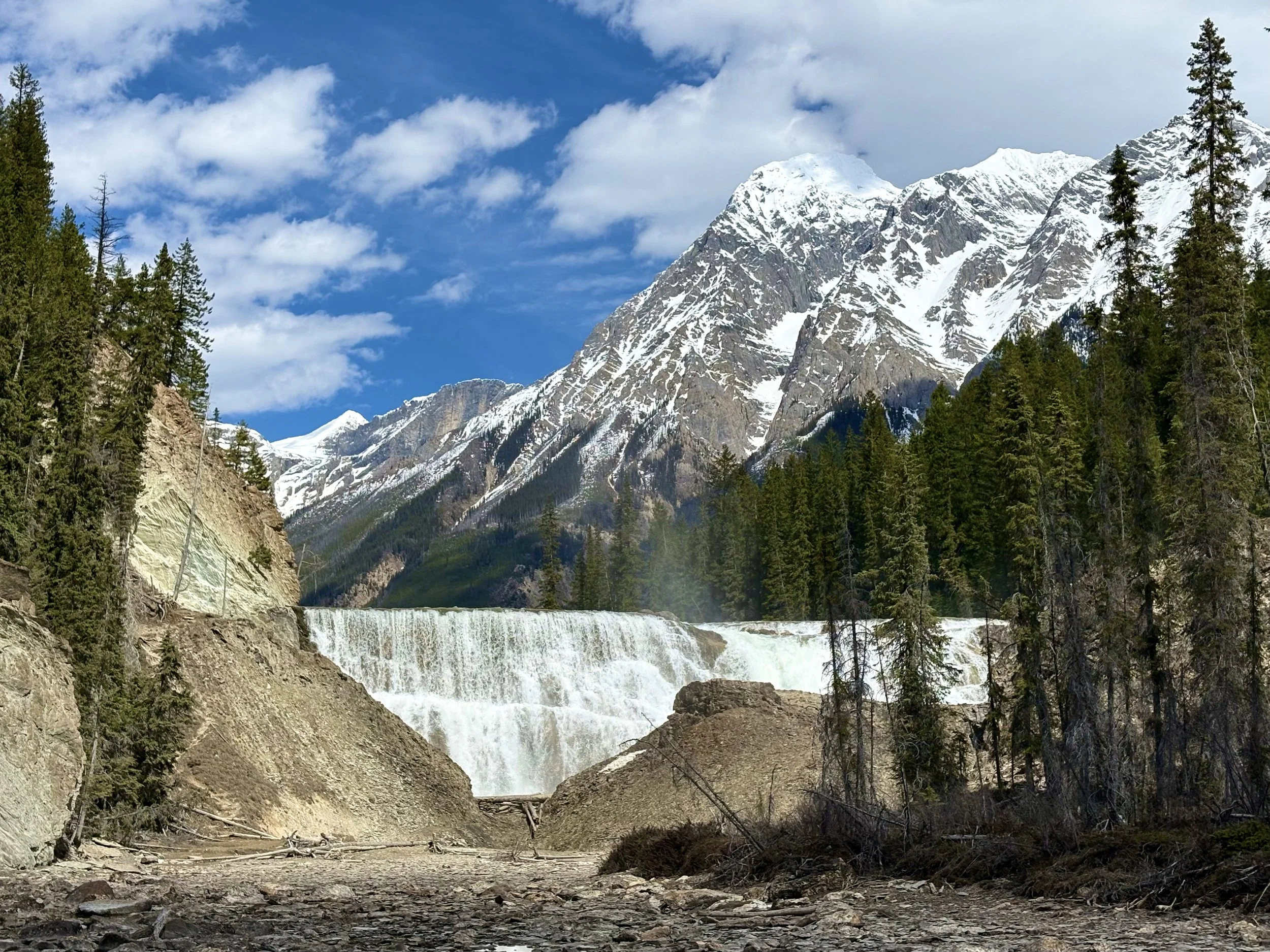 Snow-capped mountains with a blue sky and clouds, evergreen trees in the foreground, and a waterfall flowing over rocks.