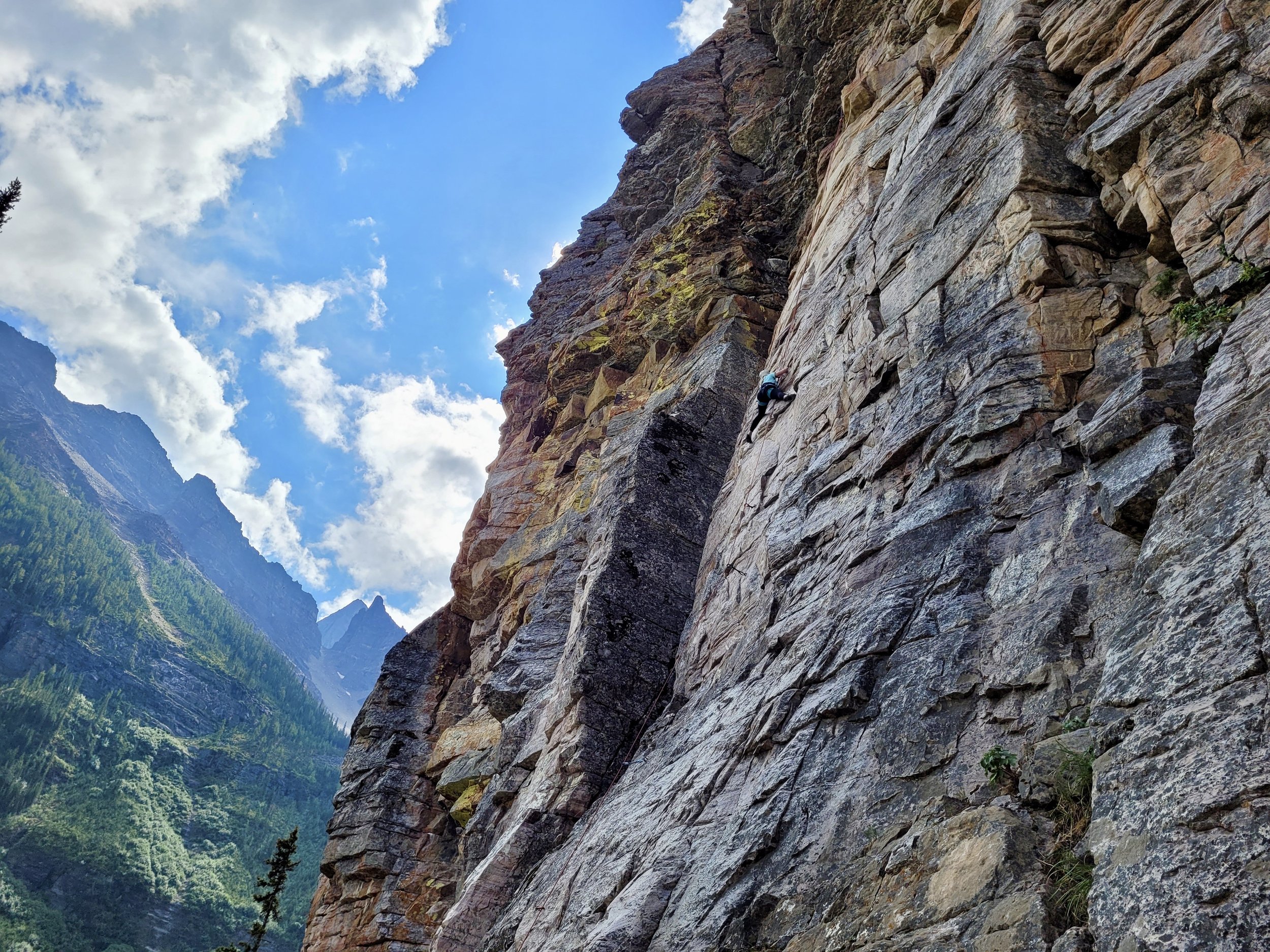 A person rock climbing on a steep, rugged cliff face in a mountainous area, with lush green trees and mountain peaks in the background under a partly cloudy sky.