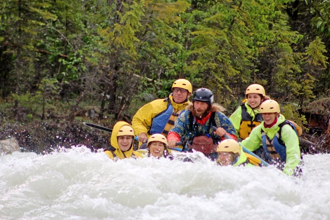 Group of six people white water rafting, wearing yellow helmets, life jackets, and rain gear, paddling through rapids with greenery in the background.