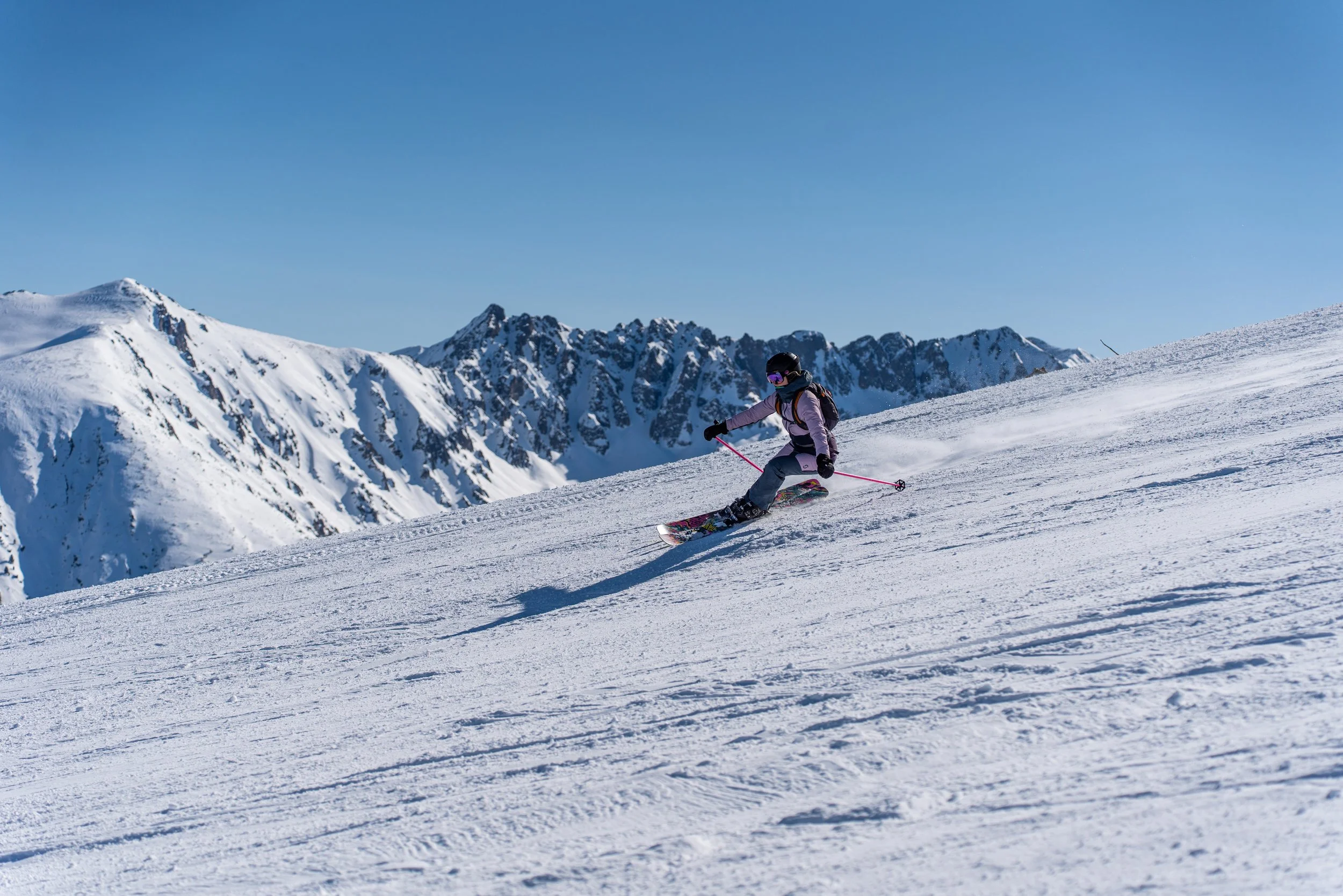 A person skiing on a snow-covered mountain slope with rugged, snow-capped peaks in the background under a clear blue sky.