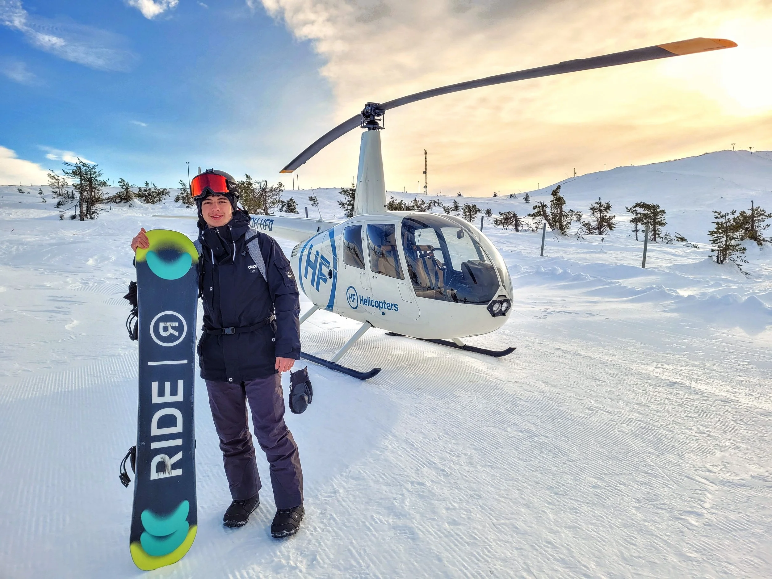 A man dressed in winter gear, holding a snowboard, standing next to a helicopter on a snowy landscape with trees and hills in the background during sunset.