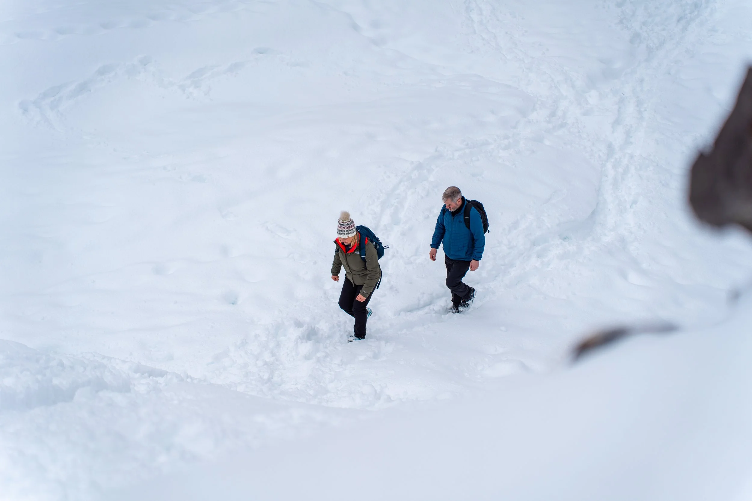 Two people are walking in snow, dressed in winter clothing with backpacks, leaving footprints in the snow.
