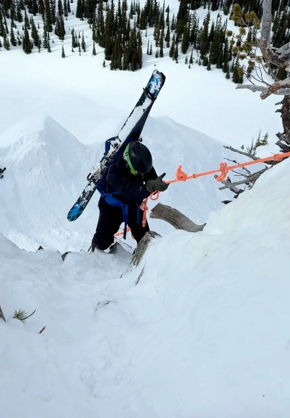 Climber in black gear ascending snowy mountain, carrying skis on shoulder, reaching for orange safety rope, surrounded by snow and trees.