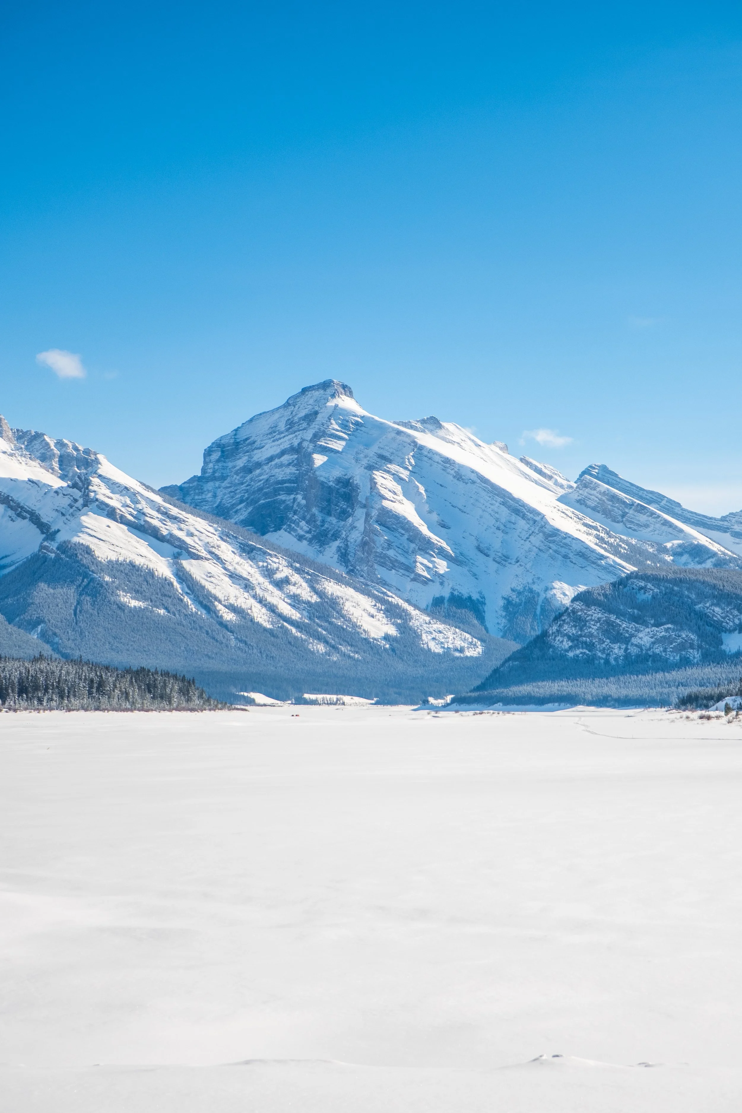 Snow-covered mountain range under a clear blue sky with a flat snowfield in the foreground.