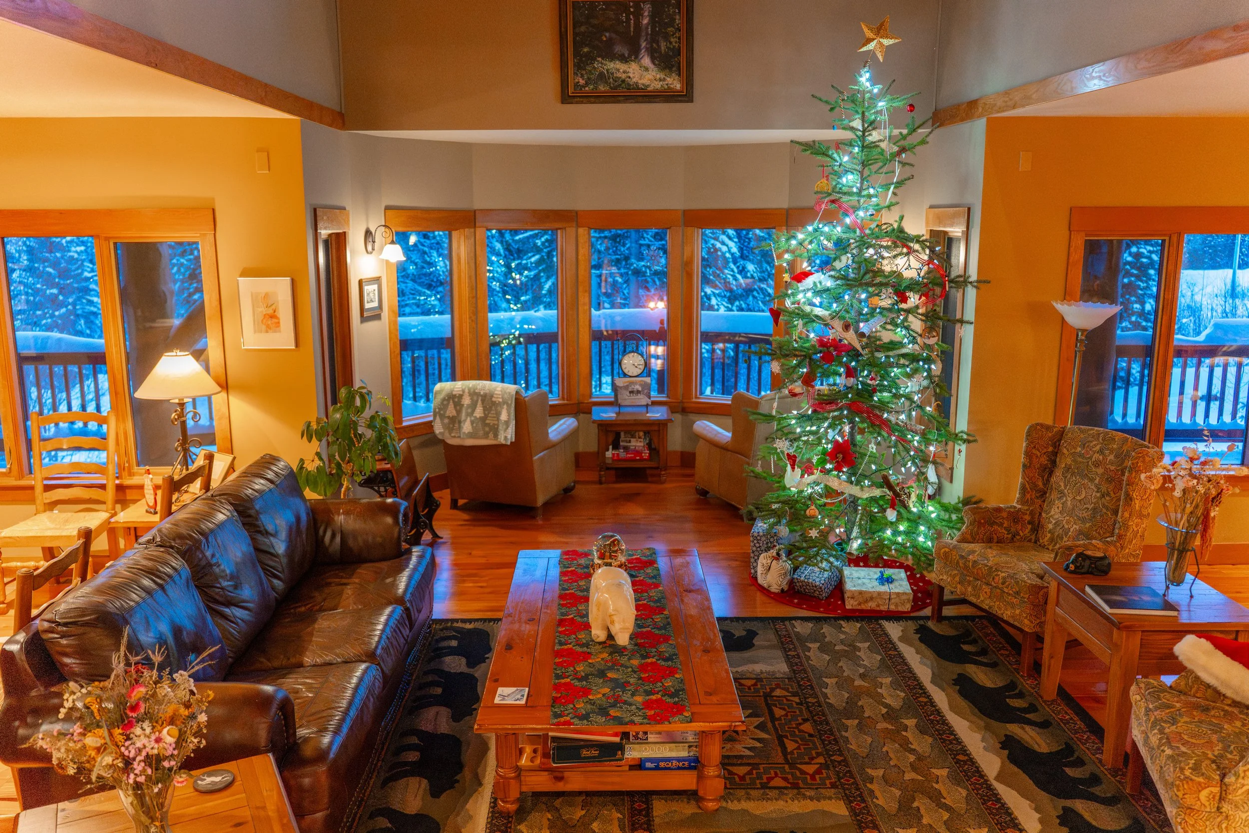 Living room decorated for Christmas with a Christmas tree, wrapped presents, and holiday decorations, with a view of snowy trees outside the windows.