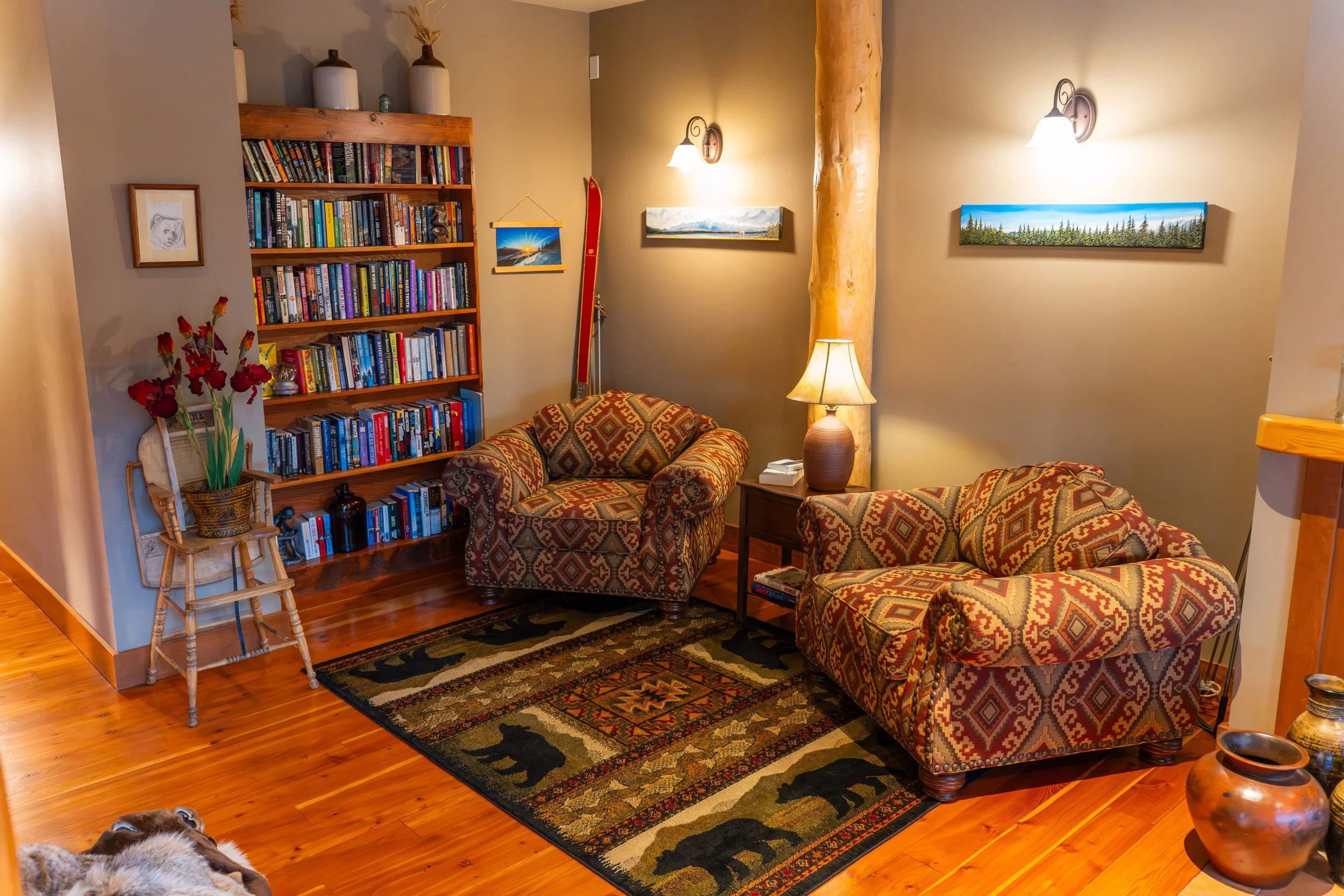 Living room with patterned armchairs, a wooden bookshelf filled with books, a table lamp, paintings on the wall, a small chair with flowers, a colorful rug with bears, and wooden flooring.