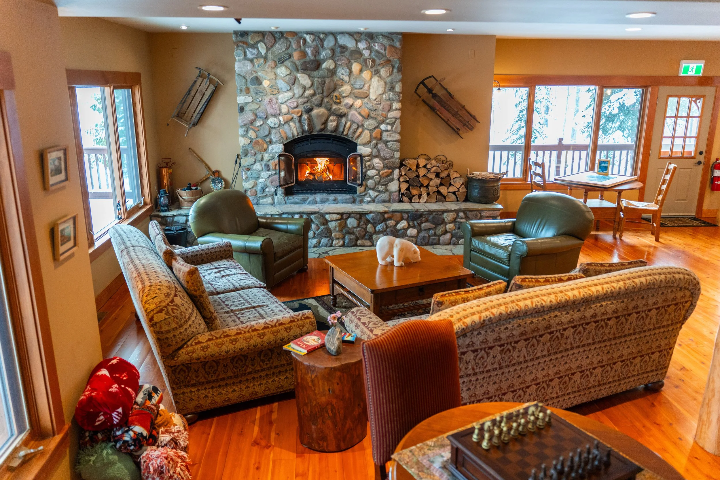 Cozy living room with a stone fireplace, leather and patterned upholstered chairs, wooden tables, and wood flooring. A fire burns in the fireplace, and wood logs are stacked nearby. Large windows and a glass door lead to a balcony with trees outside.