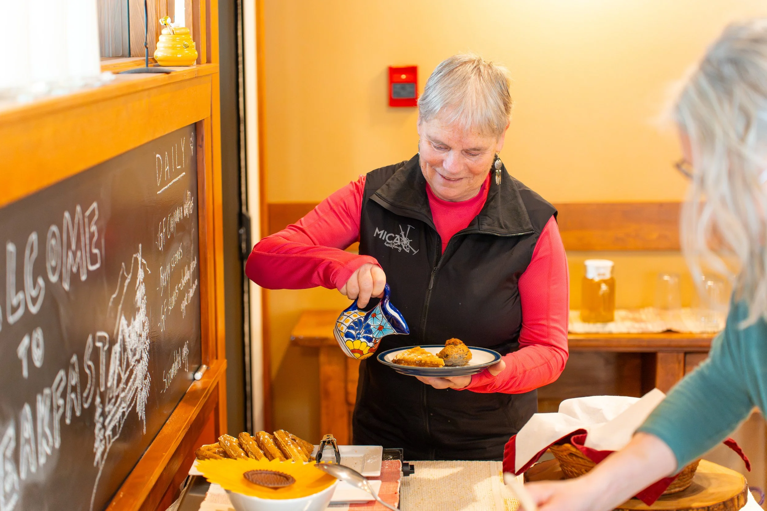 A woman serving food at a buffet table with baked goods, in a warm yellow room with a chalkboard and wooden accents.