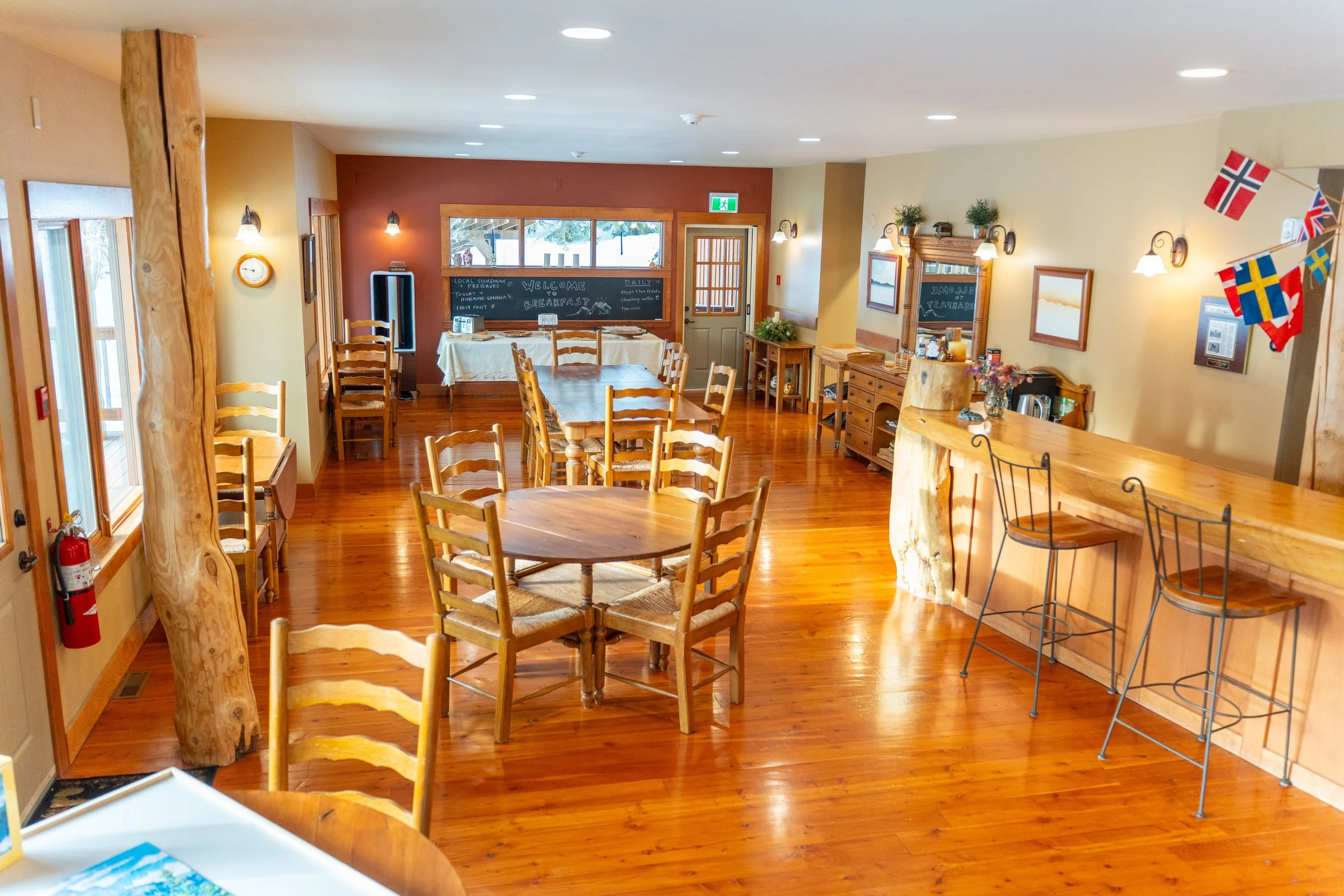 A cozy wooden interior dining area with tables and chairs, decorated with flags and paintings, featuring warm lighting and large windows showing snow outside.
