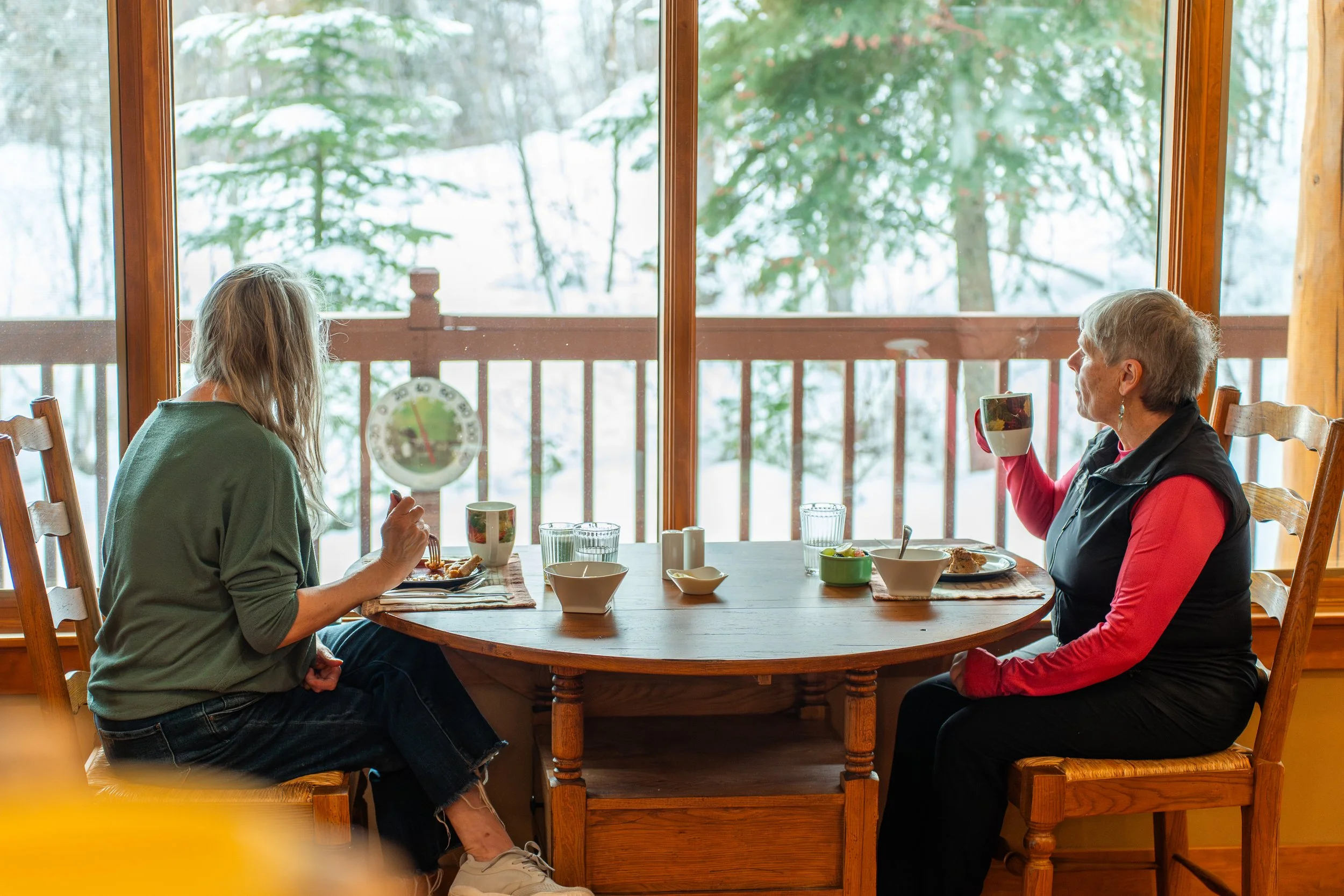 Two women sitting at a wooden dining table near large windows overlooking snowy trees, sharing a meal and coffee.