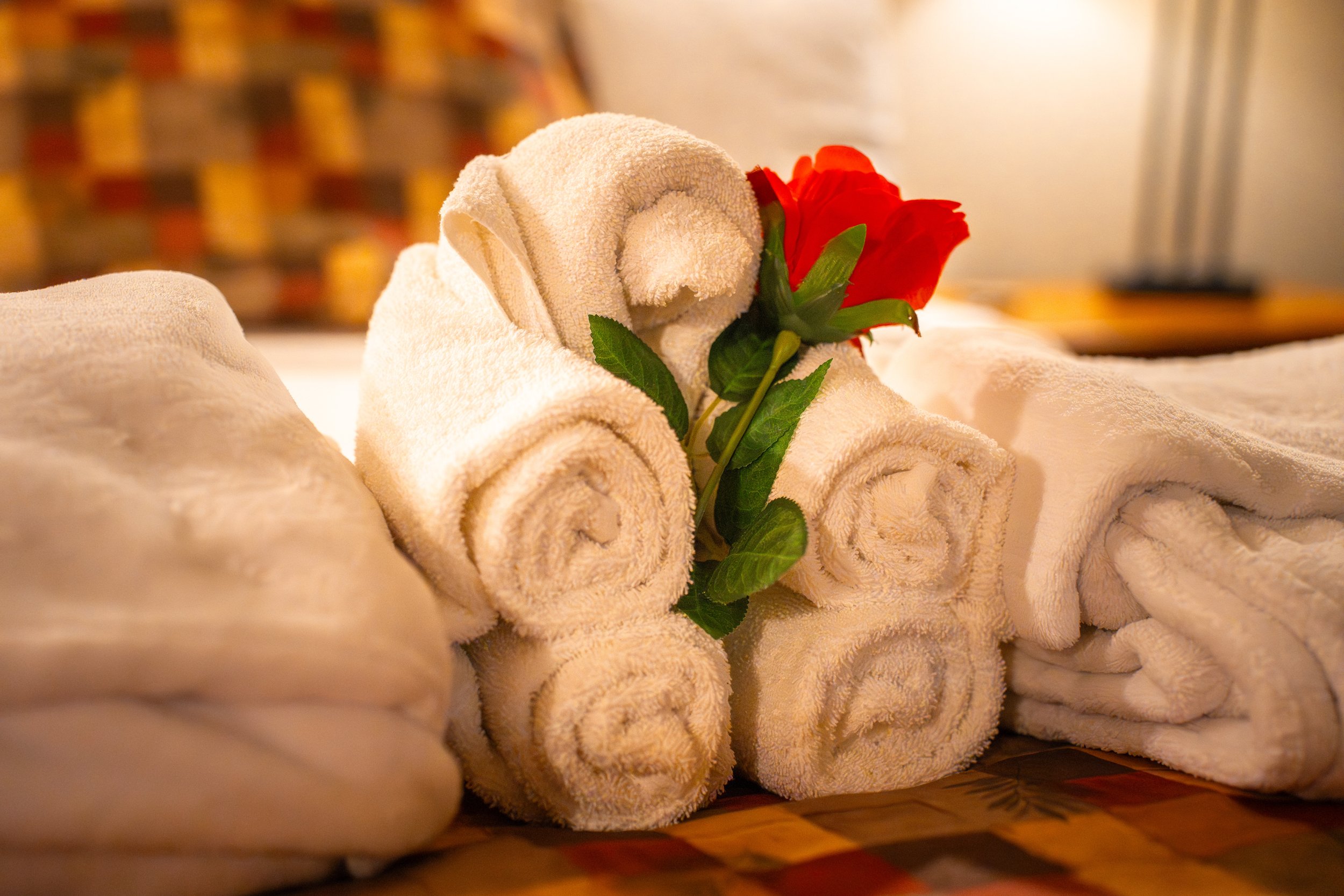 A stack of neatly rolled white towels with a red rose and green leaves on top, placed on a patterned surface.