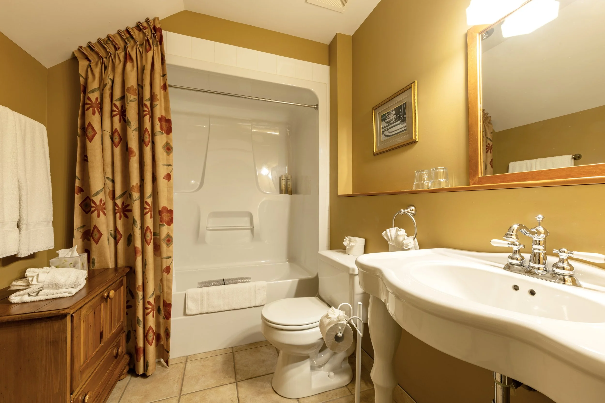 A small bathroom with a bathtub and shower with floral-patterned curtains, a wooden cabinet with towels, a toilet, a sink with a mirror, and wall decorations, illuminated by warm lighting.