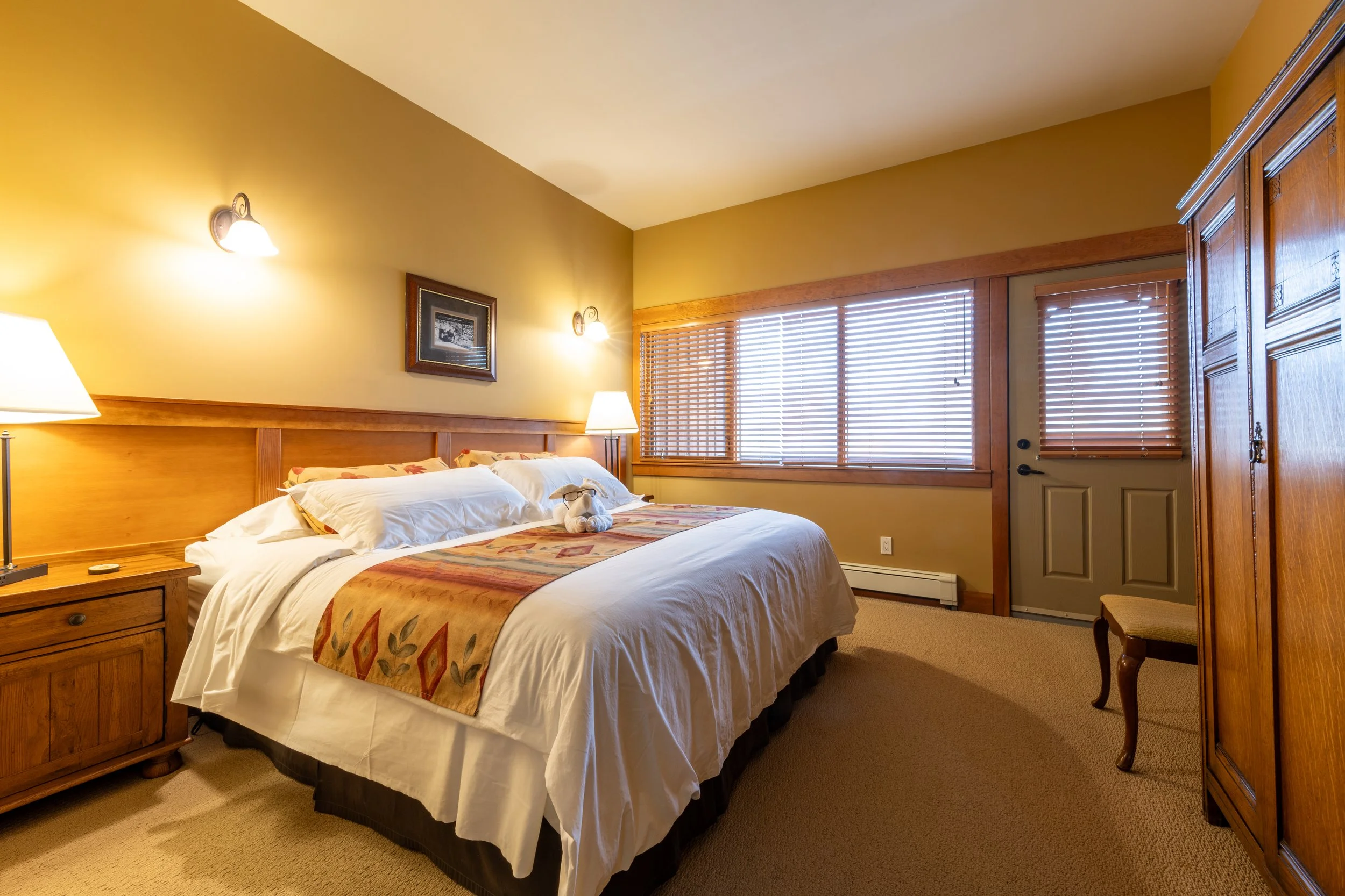 Hotel room with a bed, nightstand, lamps, framed picture, window with blinds, and wooden furniture.
