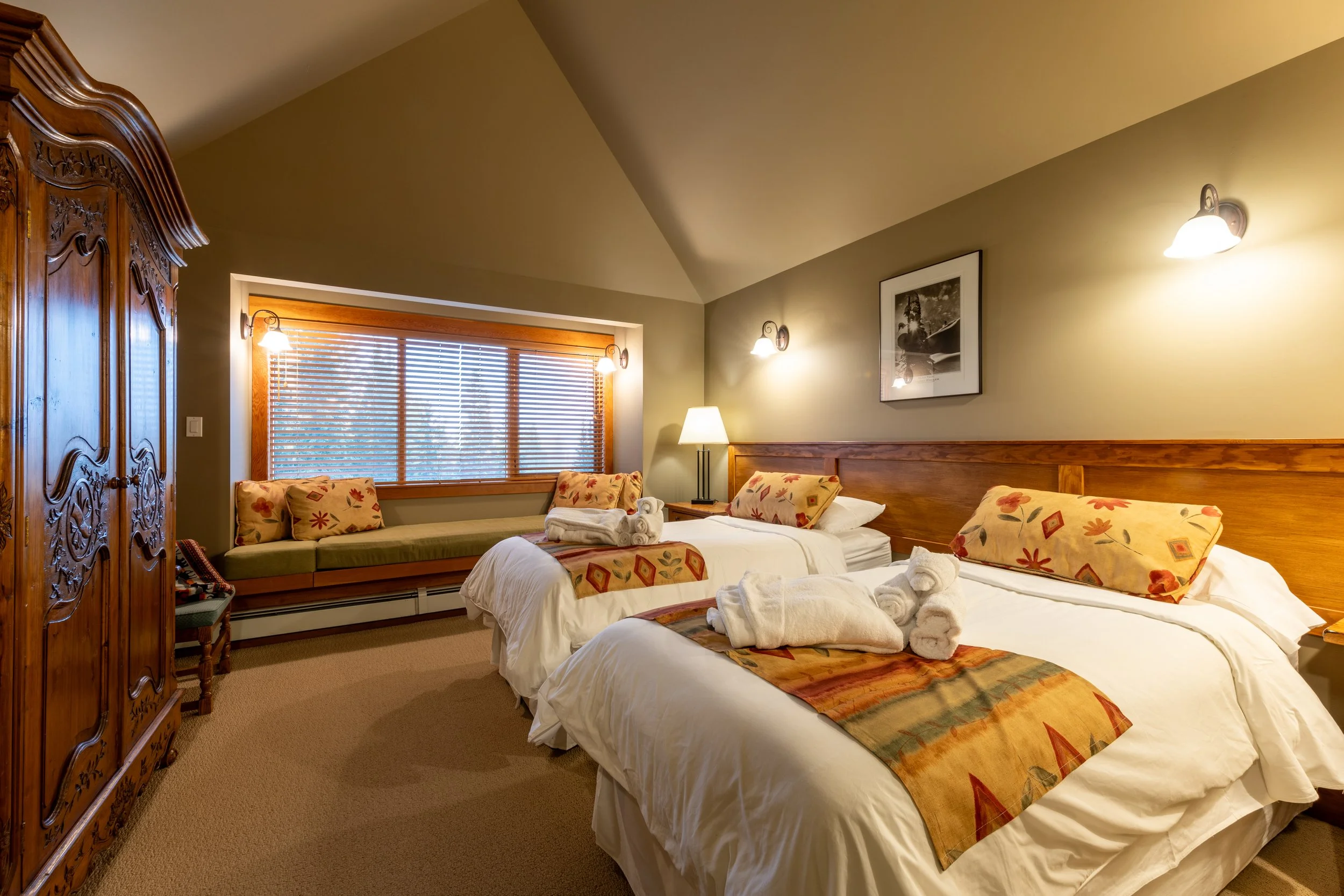 A bedroom with two beds, a wooden wardrobe, a window with blinds, and wall-mounted lights.